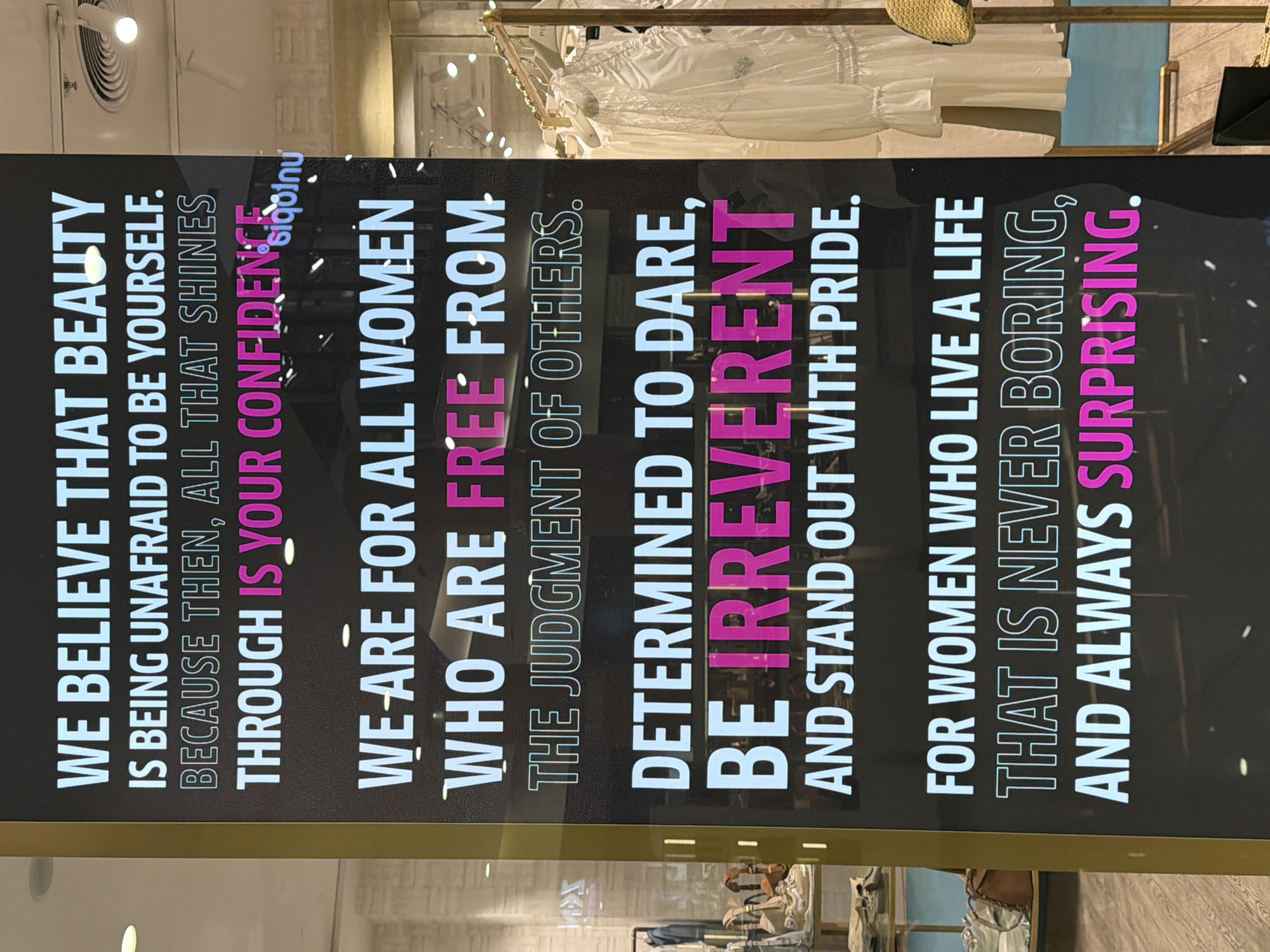 A brightly lit digital display, positioned vertically within an indoor retail environment in the city of Unknown, Unknown, serves as the central subject. The display features white and magenta text on a black background, advocating for female empowerment. The text reads: "WE BELIEVE THAT BEAUTY IS BEING UNAFRAID TO BE YOURSELF. BECAUSE THEN, ALL THAT SHINES THROUGH IS YOUR CONFIDENCE. WE ARE FOR ALL WOMEN WHO ARE FREE FROM THE JUDGMENT OF OTHERS. DETERMINED TO DARE, BE IRREVERENT AND STAND OUT WITH PRIDE. FOR WOMEN WHO LIVE A LIFE THAT IS NEVER BORING, AND ALWAYS SURPRISING." The words "CONFIDENCE," "FREE," "IRREVERENT," and "SURPRISING" are emphasized in magenta. A golden-colored vertical architectural column frames the left side of the display, adjacent to a retail window. Through this left window, reflections of overhead lights are visible, along with store merchandise including sneakers, handbags, and folded apparel displayed on shelves above a light wooden floor. To the right of the digital display, a glass storefront or open entrance reveals a clothing rack holding several white, long-sleeved dresses or blouses, one featuring lace trim. A woven straw-colored accessory, likely a hat or bag, is partially visible on the far right. Reflections of ceiling lights, including recessed spotlights and a circular air vent, are prominent on both retail windows. No individuals are present in the frame. The environment is consistent with a modern commercial shopping space.