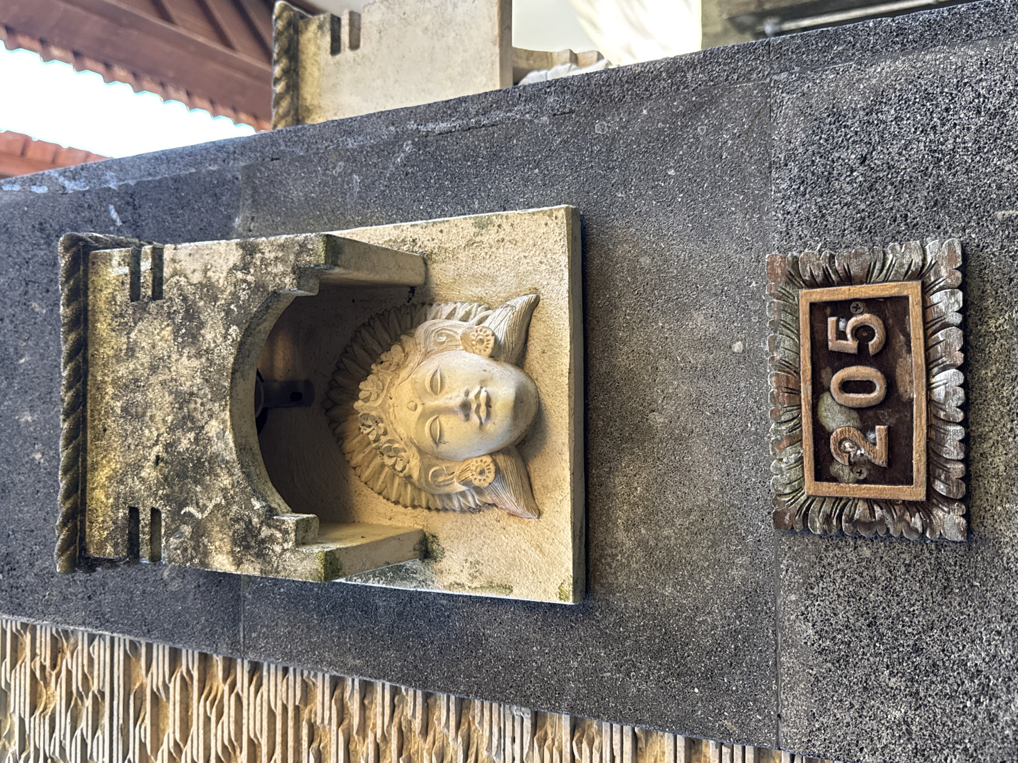 A close-up shot shows a gray stone wall in Ungasan, Indonesia. Affixed to the wall is a rectangular beige stone sculpture of a face surrounded by a textured semicircle within an open stone frame, which forms an arch over the face. To the right of the sculpture is a small, ornate wooden plaque displaying the number "205." The bottom of the wall has a vertical beige pattern, while the roof structure is visible in the background.