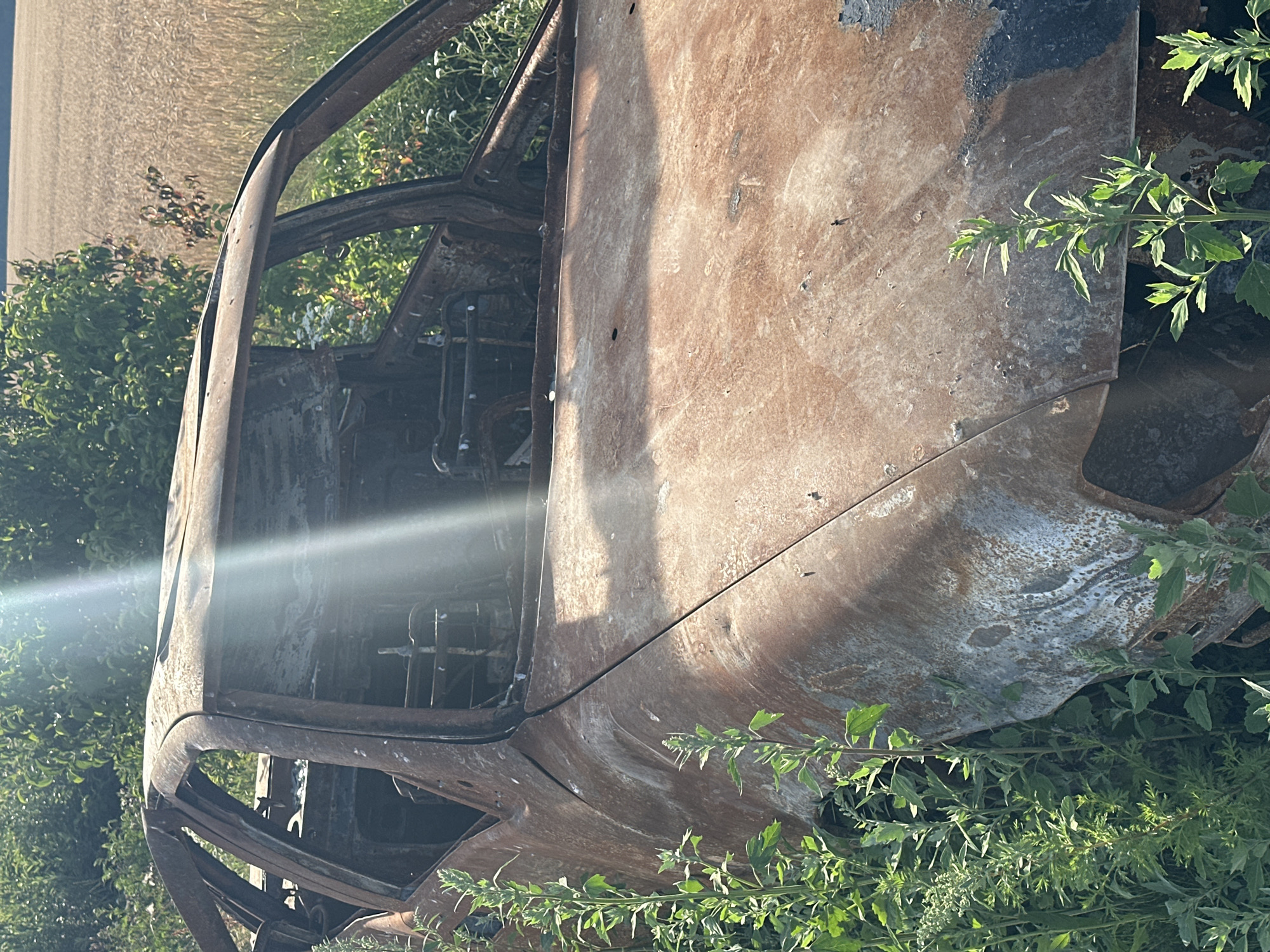 An abandoned, rusted-out car sits in a field of tall green grass and vegetation, with a wheat field visible in the background. The car appears to be a sedan, with its roof and windows missing. The interior of the car is exposed and appears to be burned out. The car is heavily damaged, with rust covering most of its exterior. The sun is shining brightly, casting a glare on the car and the surrounding vegetation. The scene suggests a rural or semi-rural location, likely in the city of Makiv, Ukraine. There are no people visible in the image.