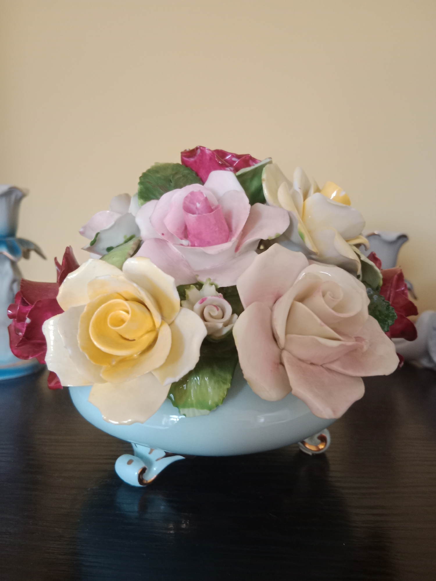 A close-up, eye-level, medium shot showcases a decorative ceramic arrangement of roses in various pastel shades of pink, yellow, and cream. The flowers are nestled in a light blue, footed bowl that rests on a dark, reflective surface, likely a tabletop. The background is a plain, light yellow wall. To the left and right of the central arrangement, out of focus, are glimpses of other decorative ceramic objects, possibly vases or figurines. The lighting is even, with no harsh shadows. The image appears to be an indoor still life, captured without any human presence. The specific location in Bydgoszcz, Poland, cannot be determined from the visual information provided.