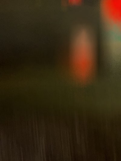 The image presents a blurred view, likely taken at night. In the upper center, there is a diffuse red glow, possibly from a light source. Below, a dark, indistinct area suggests ground cover or foliage. The bottom portion features vertical streaks, potentially indicating rain or a reflection on a wet surface. The location, Indio, United States, is not discernible from the visual information alone.