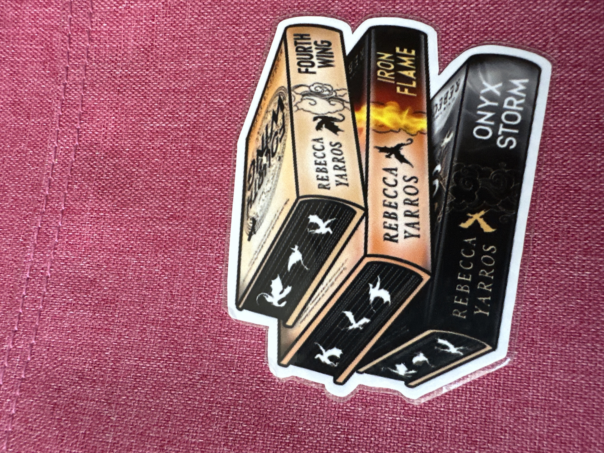 A sticker of three books, "Fourth Wing," "Iron Flame," and "Onyx Storm" by Rebecca Yarros, rests on a textured red fabric. The sticker has a white border. The environment is not identifiable as a city of Unknown, Unknown.