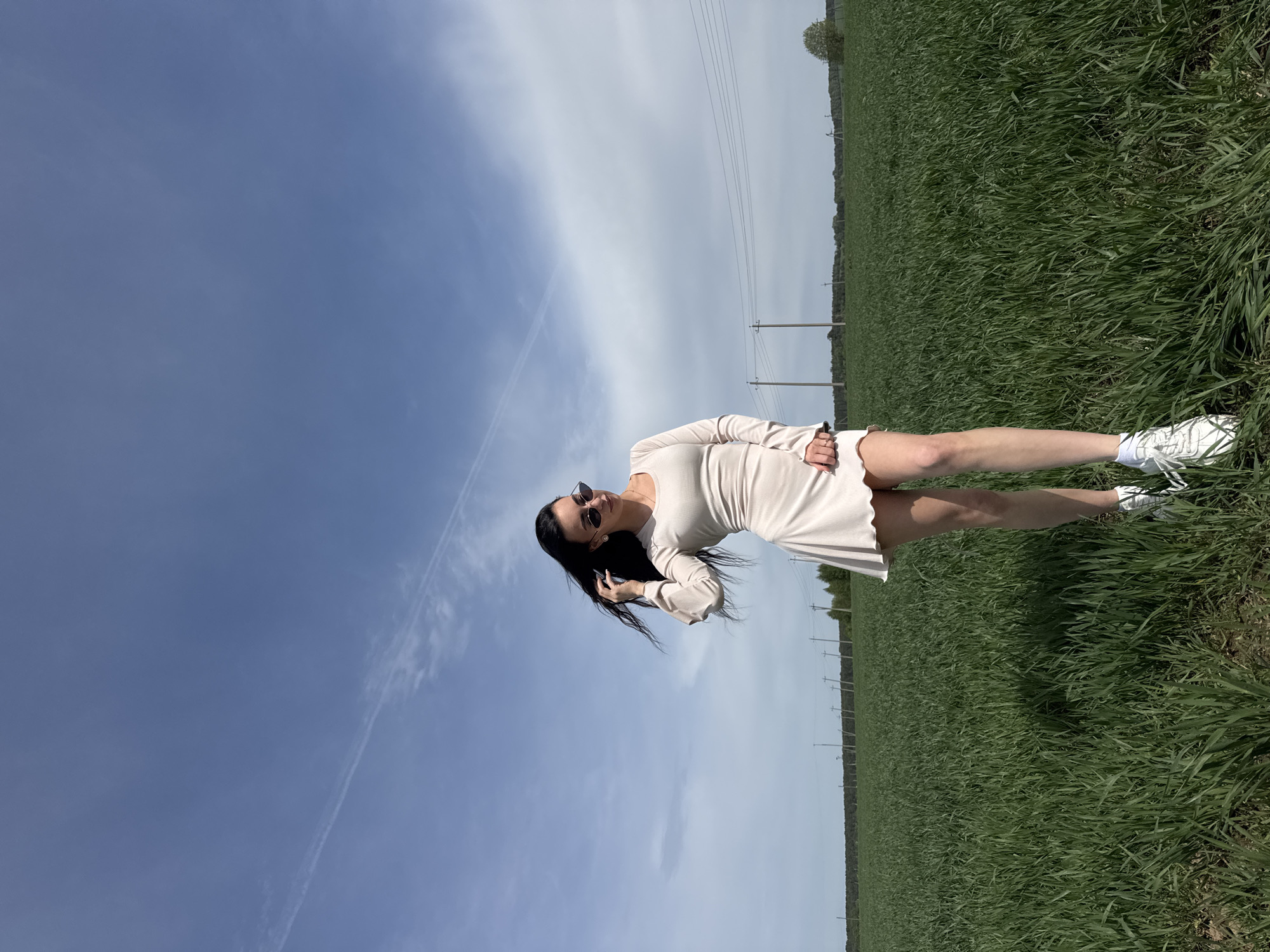 A woman stands in a field of green grass in Фоки, Russia. The sky is blue with a few clouds and power lines visible in the background. The woman is wearing a light-colored dress, sunglasses, and white sneakers. She has long, dark hair and is touching her hair with her right hand. The image appears to be rotated 90 degrees counter-clockwise.