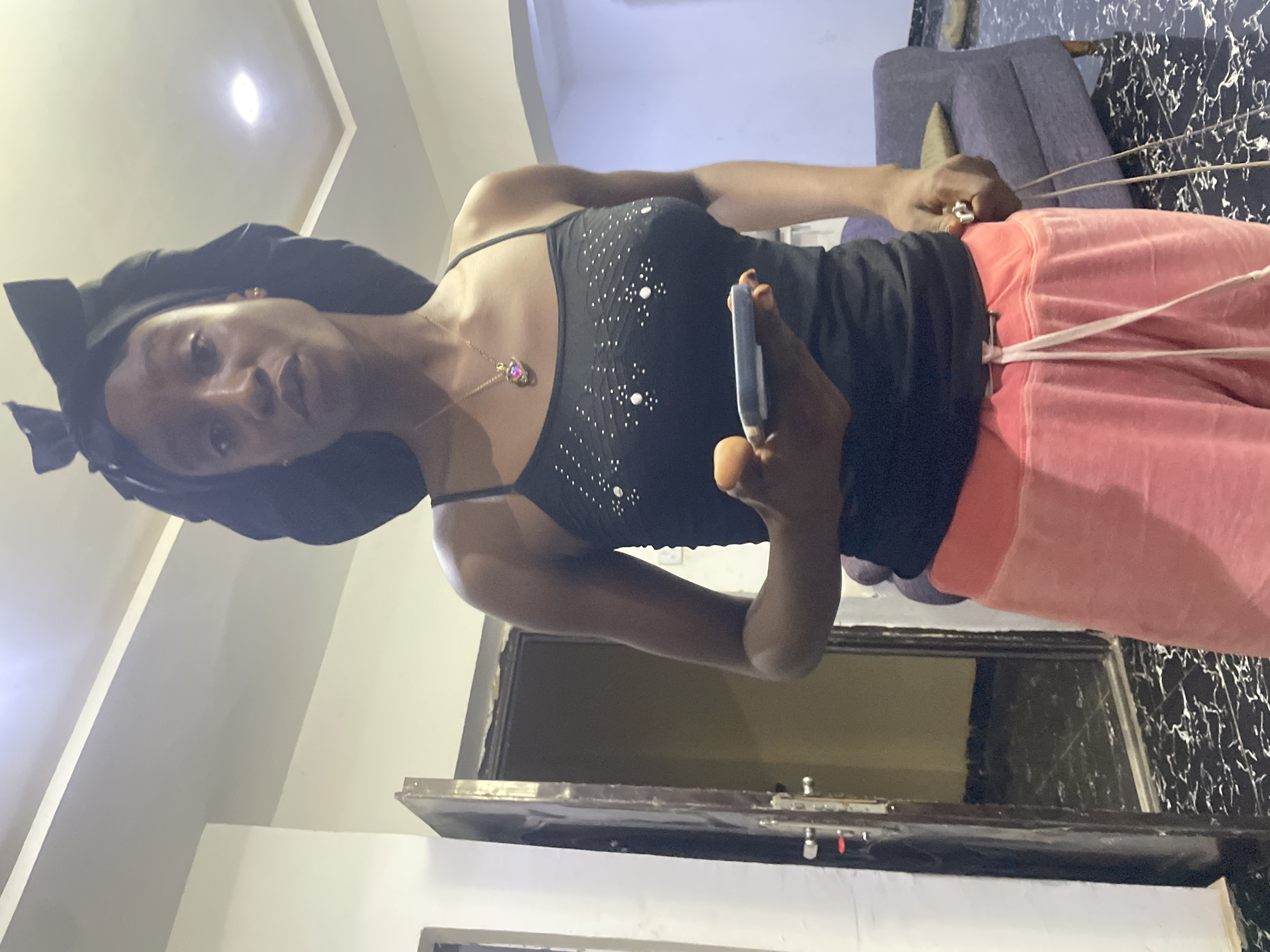 A young Black woman stands in a well-lit room, holding a mobile phone in her right hand. She is wearing a black spaghetti strap tank top adorned with white rhinestones, and loose-fitting coral pink sweatpants. A black bonnet with a large bow is on her head. A gold necklace with a heart-shaped pendant is visible around her neck. The room features white walls and a tiled floor with a black and white marble pattern. To the left, a dark brown wooden door is ajar, revealing a dimly lit interior. In the background, a purple sofa with a brown cushion is visible. The overall impression is of a casual, indoor setting, possibly a residence in Abuja, Nigeria.