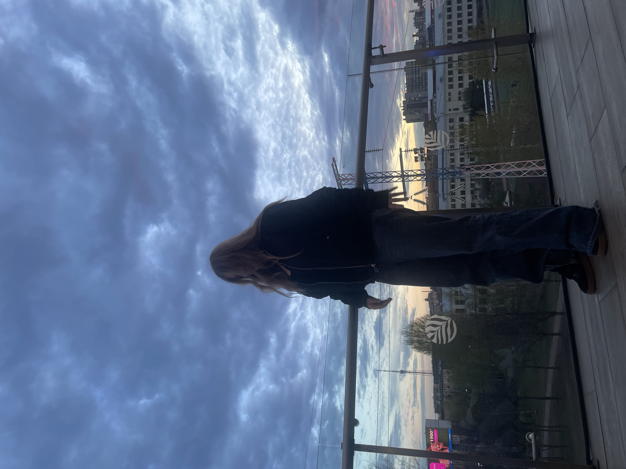 A young, long-haired person stands on a modern balcony, gazing out over the city of Saint Petersburg, Russia. The person wears a dark jacket and jeans. They stand by a glass barrier, overlooking the cityscape. In the distance, buildings and infrastructure are visible, framed by a dramatic sky with a mix of cloud cover and sunset hues. Some electrical poles and advertising billboards with Russian text are visible on the street. The location is inferred from the combination of architectural style and the presence of Russian-language signs.