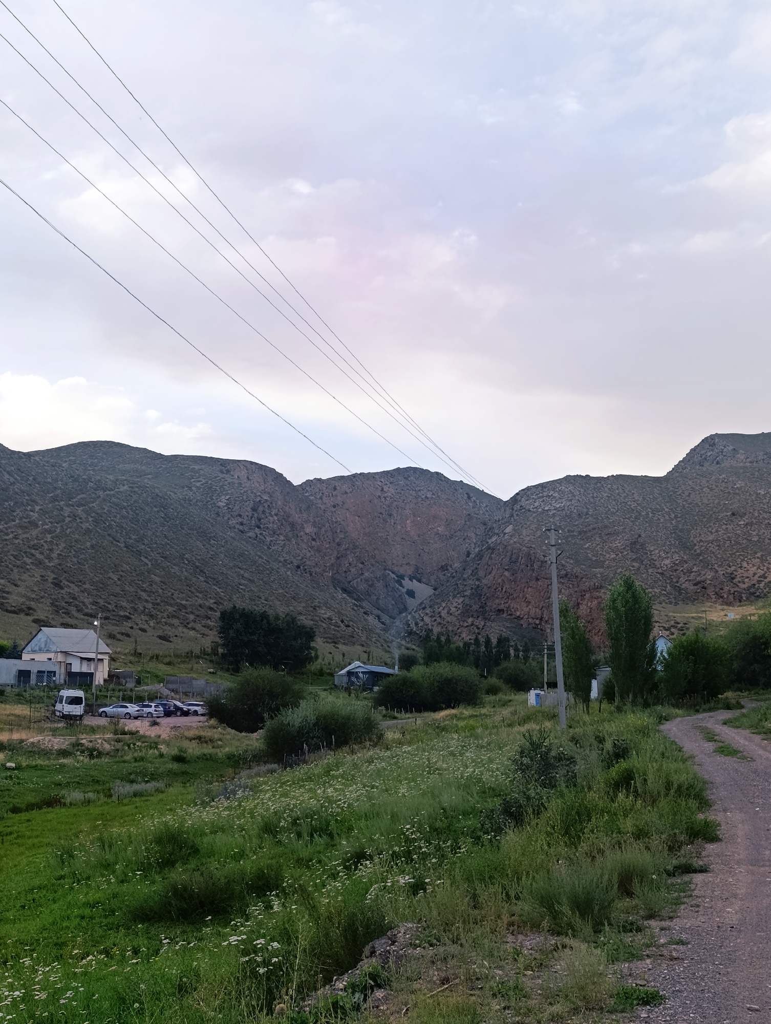 The image depicts a rural scene in Kazan-Kuygan, Kyrgyzstan, dominated by rolling hills and a cloudy sky. In the foreground, a dirt road curves to the right, bordered by green grass and wildflowers. Further back, a cluster of buildings appears, including a white house with a garage and several parked cars. Beyond the buildings, a line of trees and bushes separates the immediate area from the steep, rocky slopes of the mountains that form the backdrop. Overhead, a series of power lines stretch across the sky, crossing the scene from left to right. The overall atmosphere is calm and serene, suggesting a remote, natural setting.