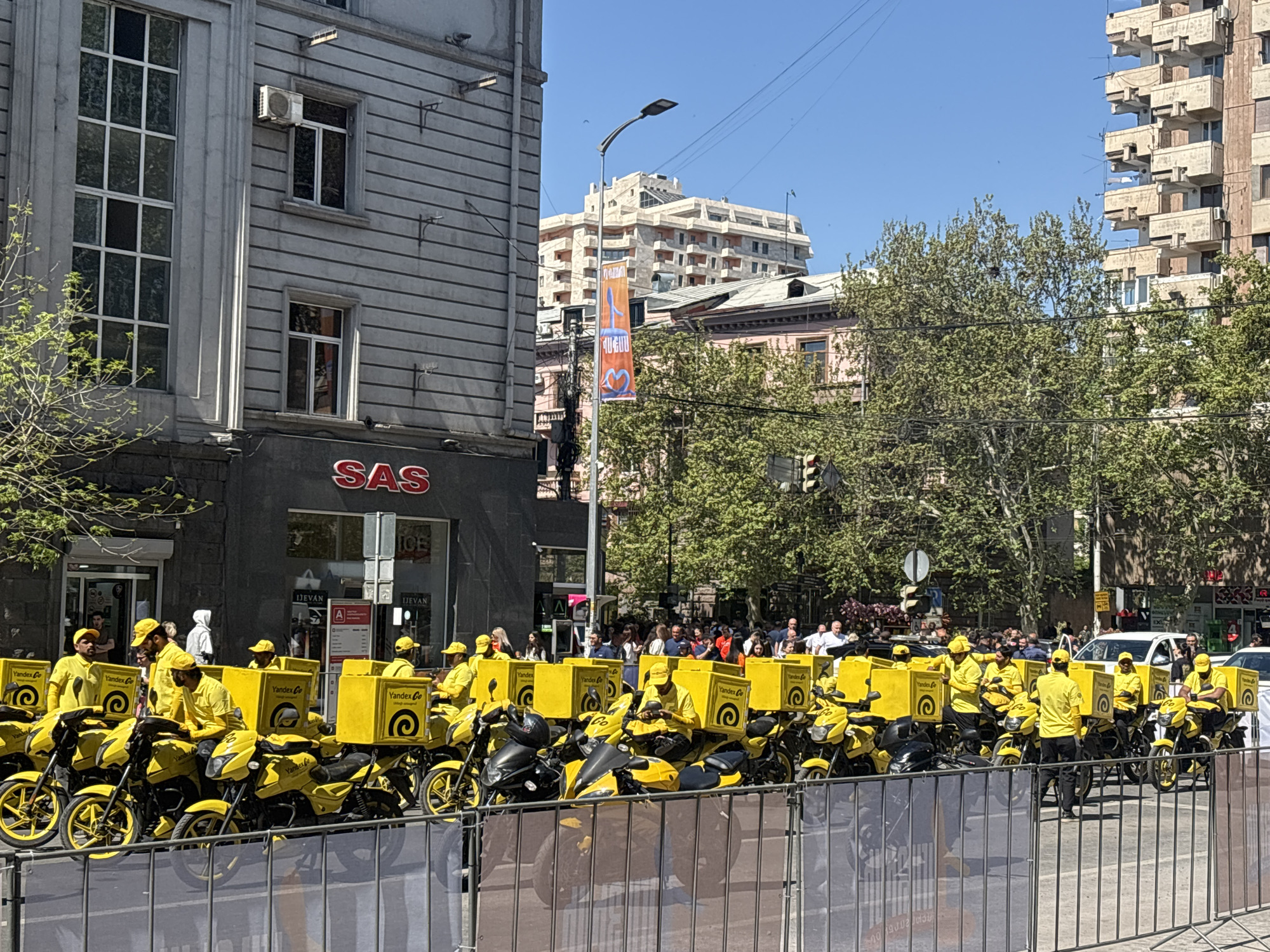 Here is a description of the image:
The image shows a row of Yandex Go delivery drivers on their mopeds in Yerevan, Armenia. The drivers are all wearing matching yellow shirts and hats, and their mopeds are also yellow with Yandex Go delivery boxes attached. The drivers are lined up behind a metal barricade, and there is a crowd of people watching them. In the background, there are several buildings, including one with a SAS sign. The sky is blue and the sun is shining.