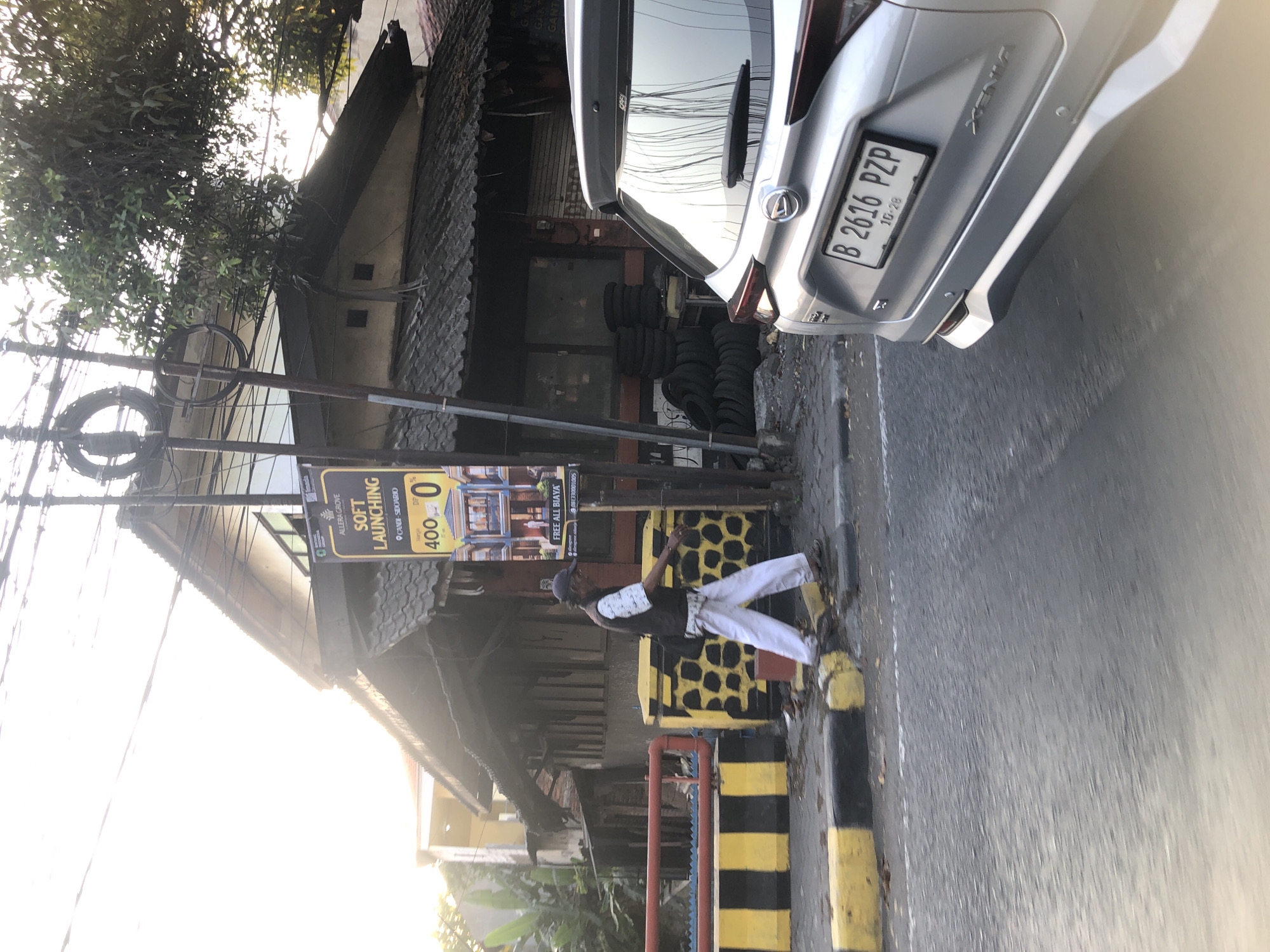 The image depicts a street scene in Larangan, Indonesia, with a silver car in the foreground and a person walking on the sidewalk. The car has a license plate that reads "B 2616 PZP". Behind the car, a person in white pants and a dark vest is walking away from the camera. In the background, there is a building with a tiled roof, a sign advertising "SOFT LAUNCHING", and several stacks of tires. The street is paved, and there are yellow and black striped barriers along the sidewalk. The sun is setting, casting a warm light on the scene.