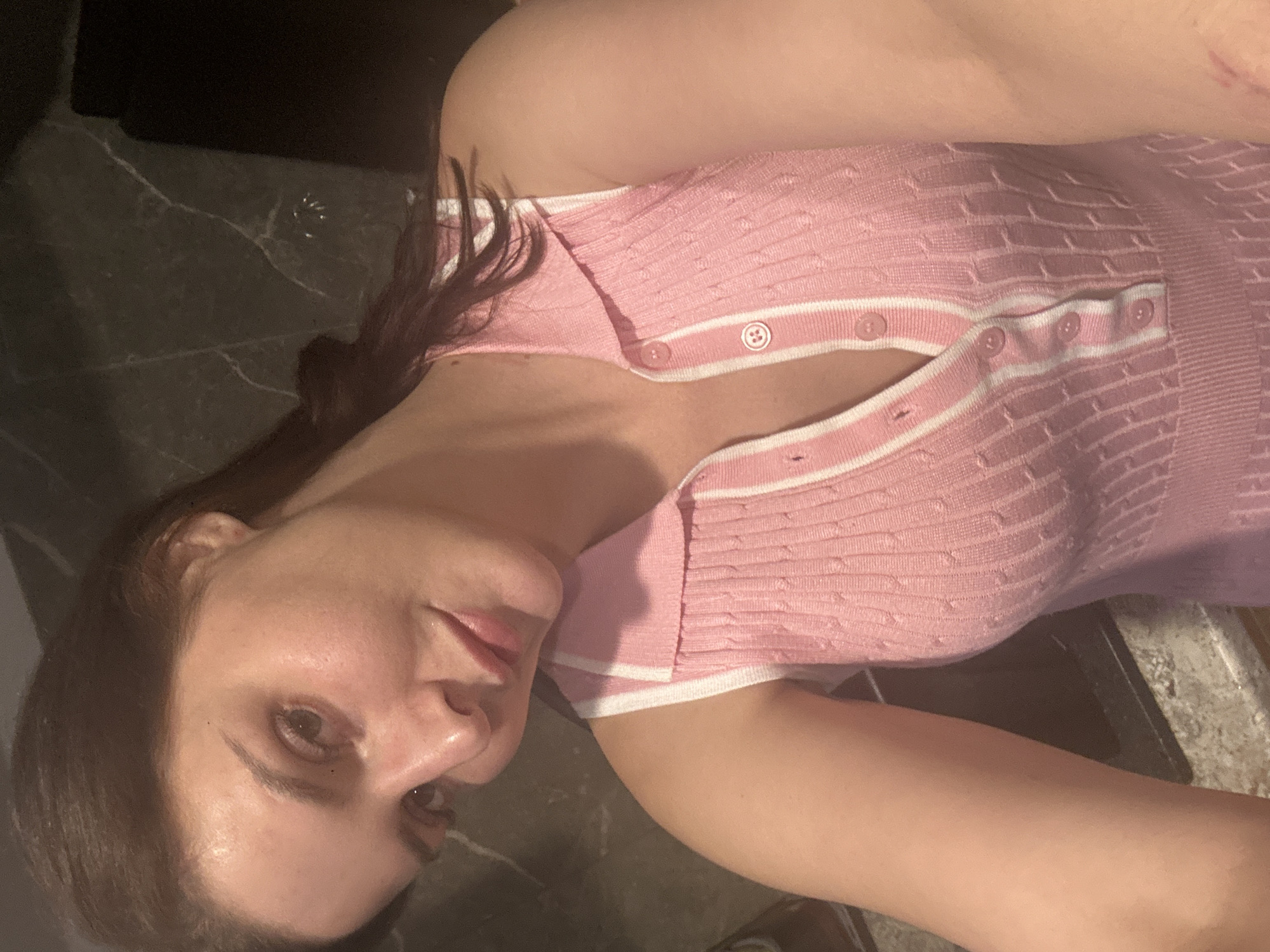An overhead, close-up shot shows a young woman with brown hair, wearing a pink collared sleeveless shirt with white trim, with a soft, slightly smiling expression. The background appears to be a dark, textured countertop, possibly marble. The woman's face is well-lit, and the lighting casts subtle shadows, highlighting her features. The image is framed to focus on her upper body and face.