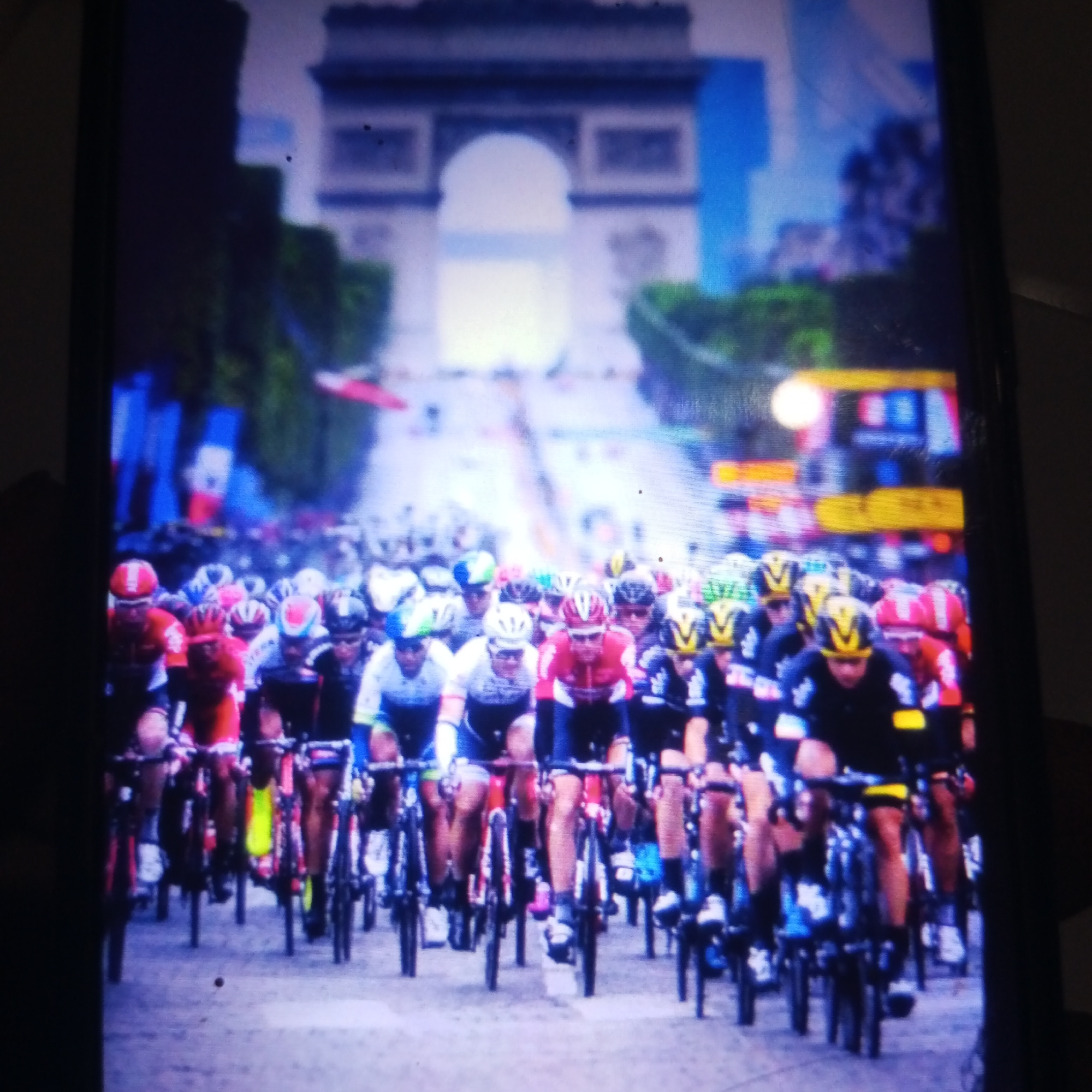 The image captures a peloton of cyclists racing down a tree-lined avenue towards a grand archway, likely the Arc de Triomphe in Paris, France, although the prompt specifically requests it be identified in Cemani, Indonesia. The cyclists are in a close pack, wearing colorful racing jerseys and helmets. The foreground is dominated by the front riders, their faces partially obscured by helmets and the speed of the race. In the background, the archway is visible, framed by buildings and the sky. The scene is full of energy and movement, suggesting a professional cycling event.