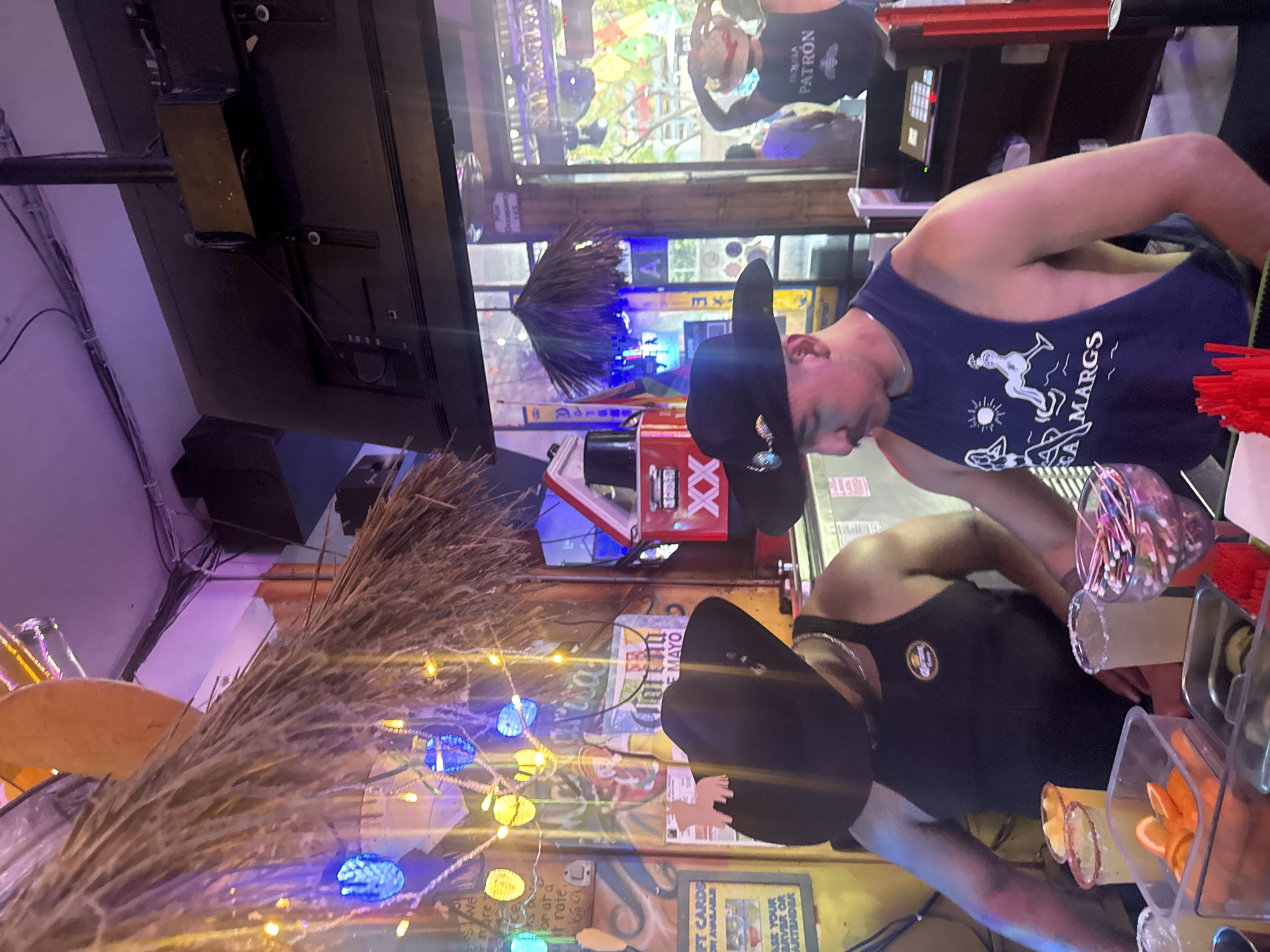 The image captures a bar scene, likely in the city of West Hollywood, United States of America. Two bartenders wearing cowboy hats and tank tops are shown preparing drinks behind the bar. One of the tank tops is labeled "MARGS" and has a graphic with a silhouette of people, a sun, and other symbols. Above them, a television screen displays an event, while a tiki-style decoration hangs near the ceiling. The bar is adorned with strings of lights, signs, and displays of beverage products such as Modelo Especial and Dos Equis. Bar tools, garnishes, and drinking glasses are visible on the bar top.
