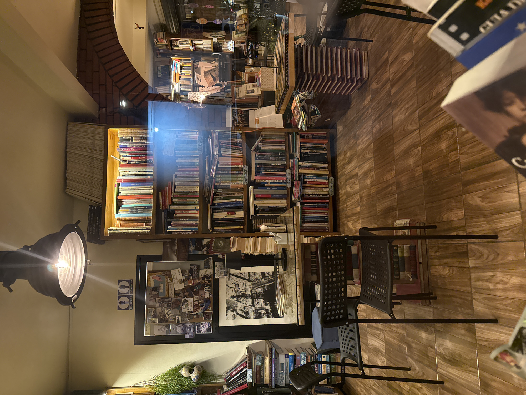The image captures the interior of Profetica Casa de la Lectura, a bookstore and cultural center located in San Andrés Cholula, Mexico. The scene is densely packed with books arranged on shelves, creating an immersive, literary environment. The lighting is warm, with a spotlight fixture illuminating the area around the shelves.
The bookstore is filled with chairs and tables, suggesting it is designed as a place for reading and interaction. Posters and photographs decorate the walls, adding to the atmosphere. A tiled floor and brickwork visible in the background add character.
Reflections in glass surfaces throughout the image create a layered effect, adding depth and complexity to the composition. The shelves hold a variety of books, with visible titles such as "Vida Americana" and "El Circulo de la Vida". The restroom signs are visible, indicating the facilities. The interior design suggests a focus on culture and community engagement.