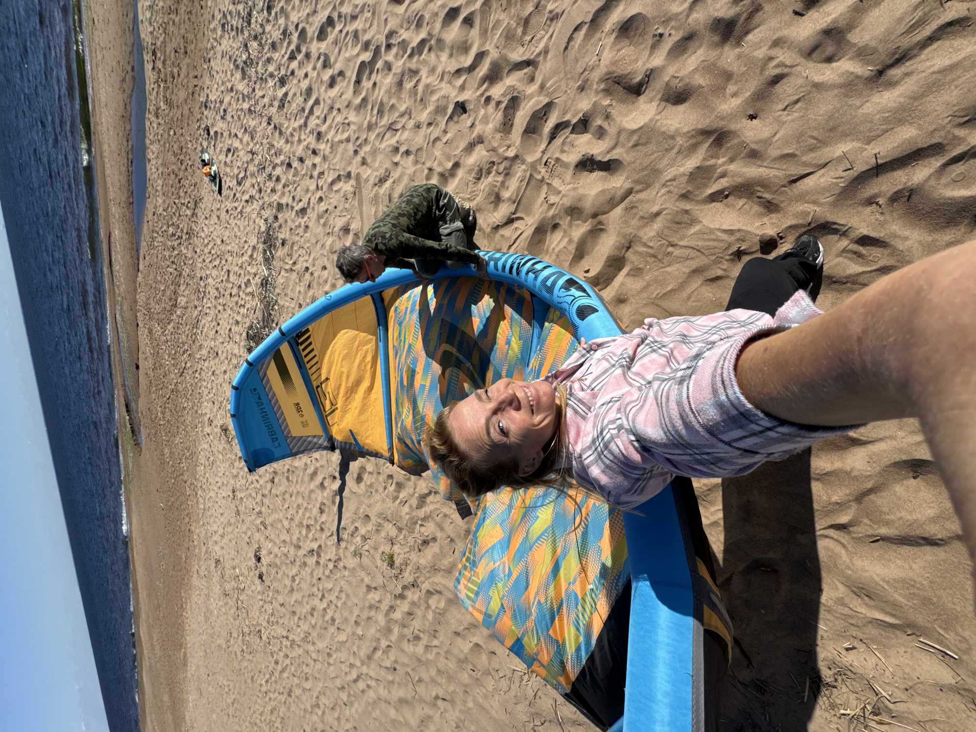 In Zelenaya Roshcha, Russia, two individuals are on a sandy beach near the Baltic Sea. A woman in a pink plaid shirt is in the foreground, holding the camera and smiling. Next to her, a large, blue and yellow kite lies on the sand. A second person, wearing a camouflage shirt and green pants, is crouched beside the kite, appearing to be working on it. The sea is visible in the background under a clear blue sky.