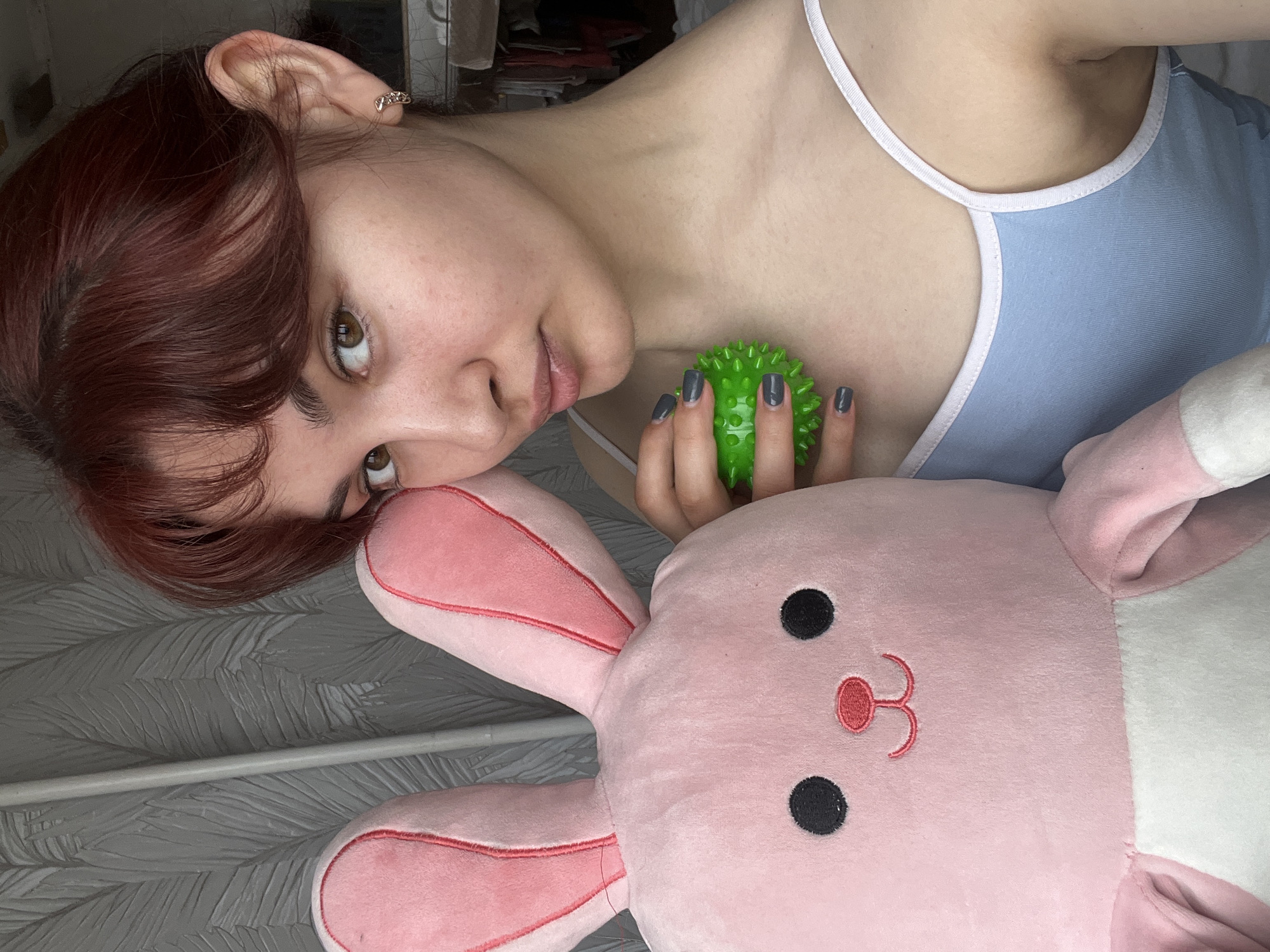 A young woman with auburn hair lies on a gray patterned surface, next to a large, pink and white stuffed rabbit. She is wearing a light blue tank top with white trim, and holding a green spiky ball in her hand. Her fingernails are painted a dark gray. The background is blurry, but there appears to be a shelf with items on it. There is no information to suggest the location of Neftekamsk, Russia in the image.