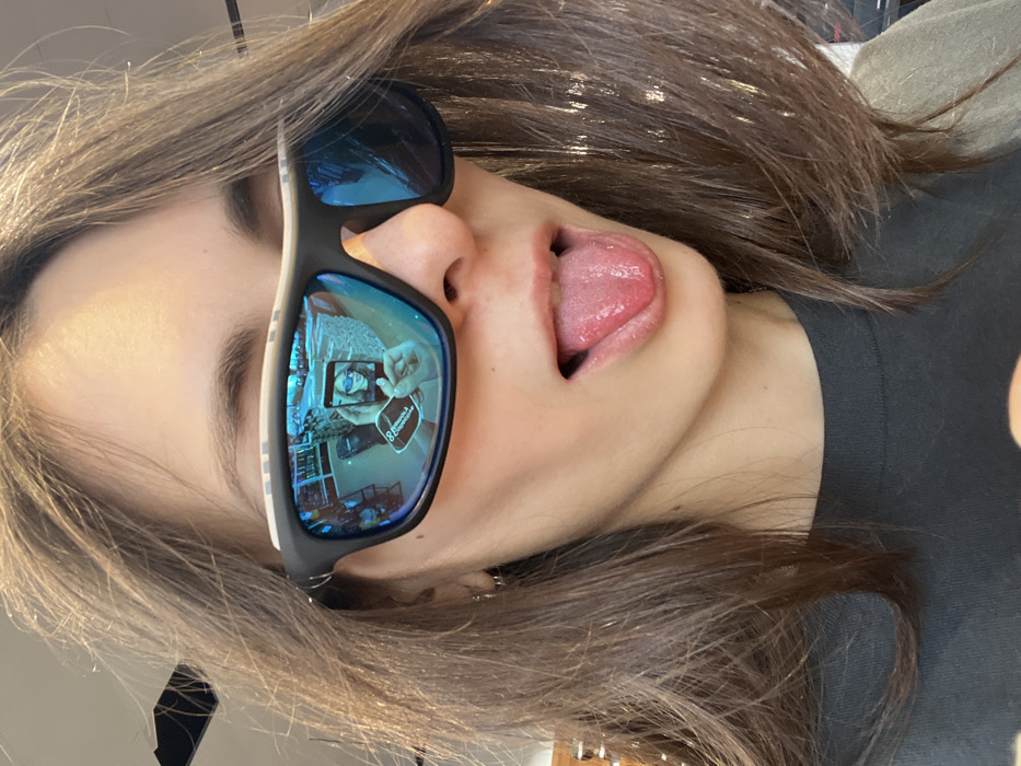 A close-up image depicts a female individual with long, brown hair, wearing black-framed sunglasses with blue reflective lenses and a dark-colored top. Her mouth is open, and her tongue is extended. The left lens of the sunglasses (viewer's left) reflects a smartphone held by her hand, displaying a selfie of the same individual. A black label or sticker on the reflected smartphone clearly displays the text "КРАСНОЯРСК & КОМПАНИЯ" (Krasnoyarsk & Company) alongside a stylized "88" logo. The background visible in the lens reflection indicates an indoor retail or shop environment with shelving and artificial lighting. This suggests the image was captured in a commercial location in Krasnoyarsk, Russia.