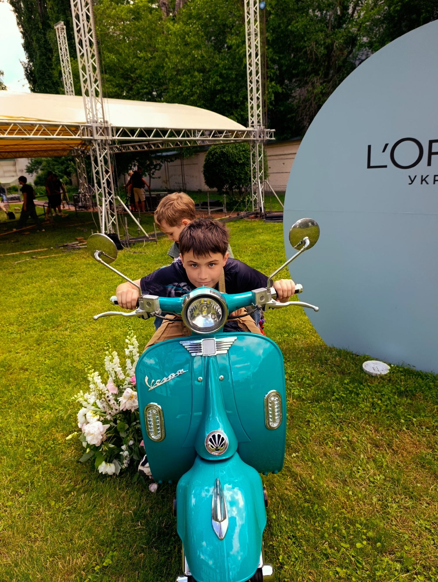 This is a medium shot, eye-level, outdoor photograph of two young boys on a teal-colored Vespa scooter on a grassy area, possibly a park or event venue in Kyiv, Ukraine. The boy in the foreground, facing the camera, is wearing a dark t-shirt and looking directly at the viewer with a serious expression. He has his hands on the scooter's handlebars. Behind him, partially obscured, is another boy with lighter hair, also looking away from the camera. The scooter is a vintage-style model with chrome accents and a prominent headlight. To the left of the scooter is a large bouquet of white and pink flowers. In the background, there is a large, light blue semi-circular structure with the L'Oréal logo and "УКР" (likely abbreviating Ukraine) printed on it in black. Further in the background, a yellow tent with metal scaffolding is visible, suggesting an outdoor event or performance setup. The ground is covered in green grass. The overall impression is of a casual, possibly promotional event.