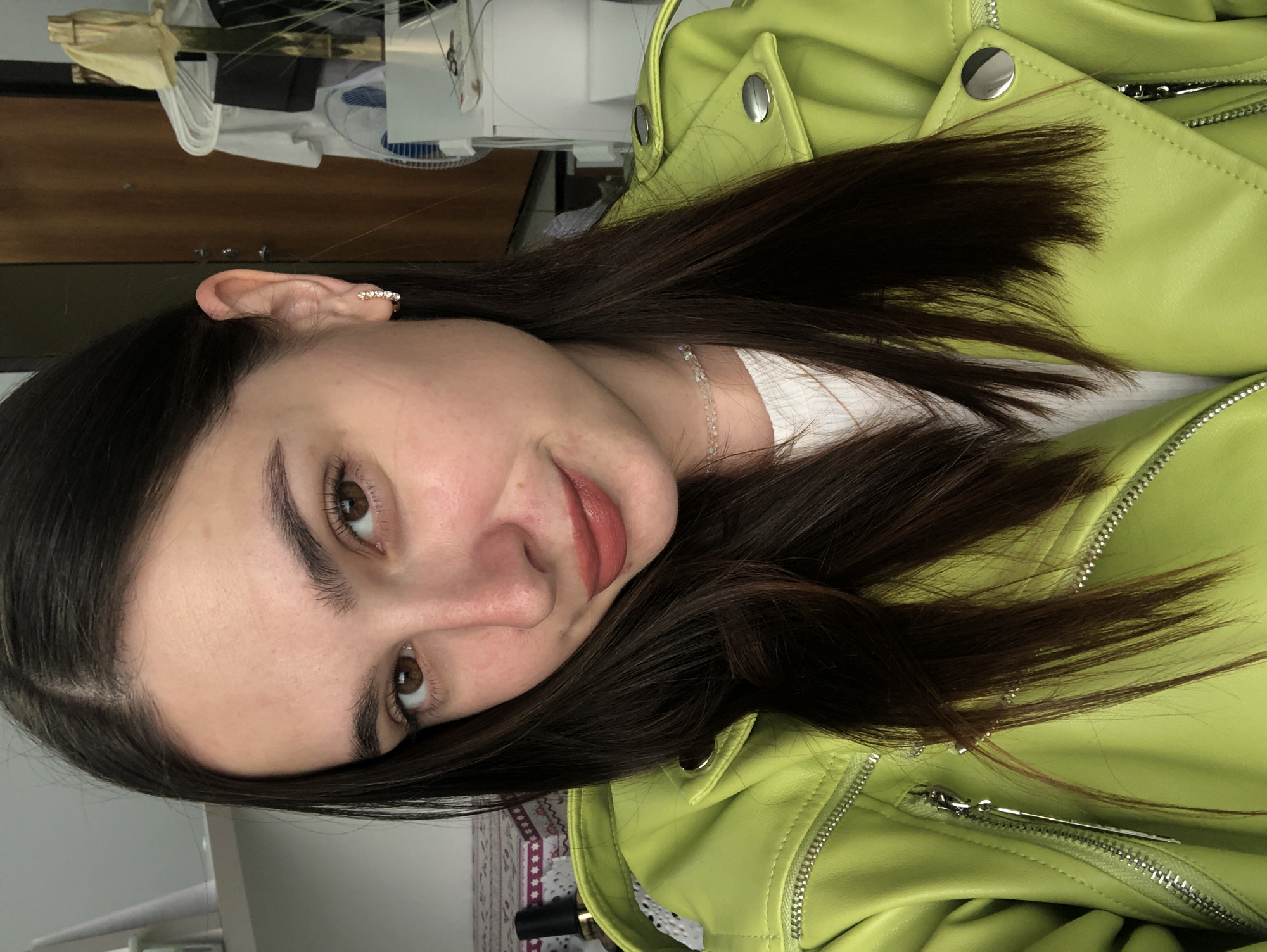 This is a vertical, medium shot of a young woman in Chaikovsky, Russia. She has long, dark brown hair and is wearing a bright lime green leather jacket over a white top. A delicate, beaded necklace is visible around her neck. Her left ear, adorned with a small, sparkly earring, is prominent in the frame. She is looking towards the camera with a soft smile and her eyes are a warm brown. The background is slightly out of focus, showing a blurred interior with hints of furniture and what appears to be a fan. The lighting suggests it is daytime. The overall mood is casual and relaxed.