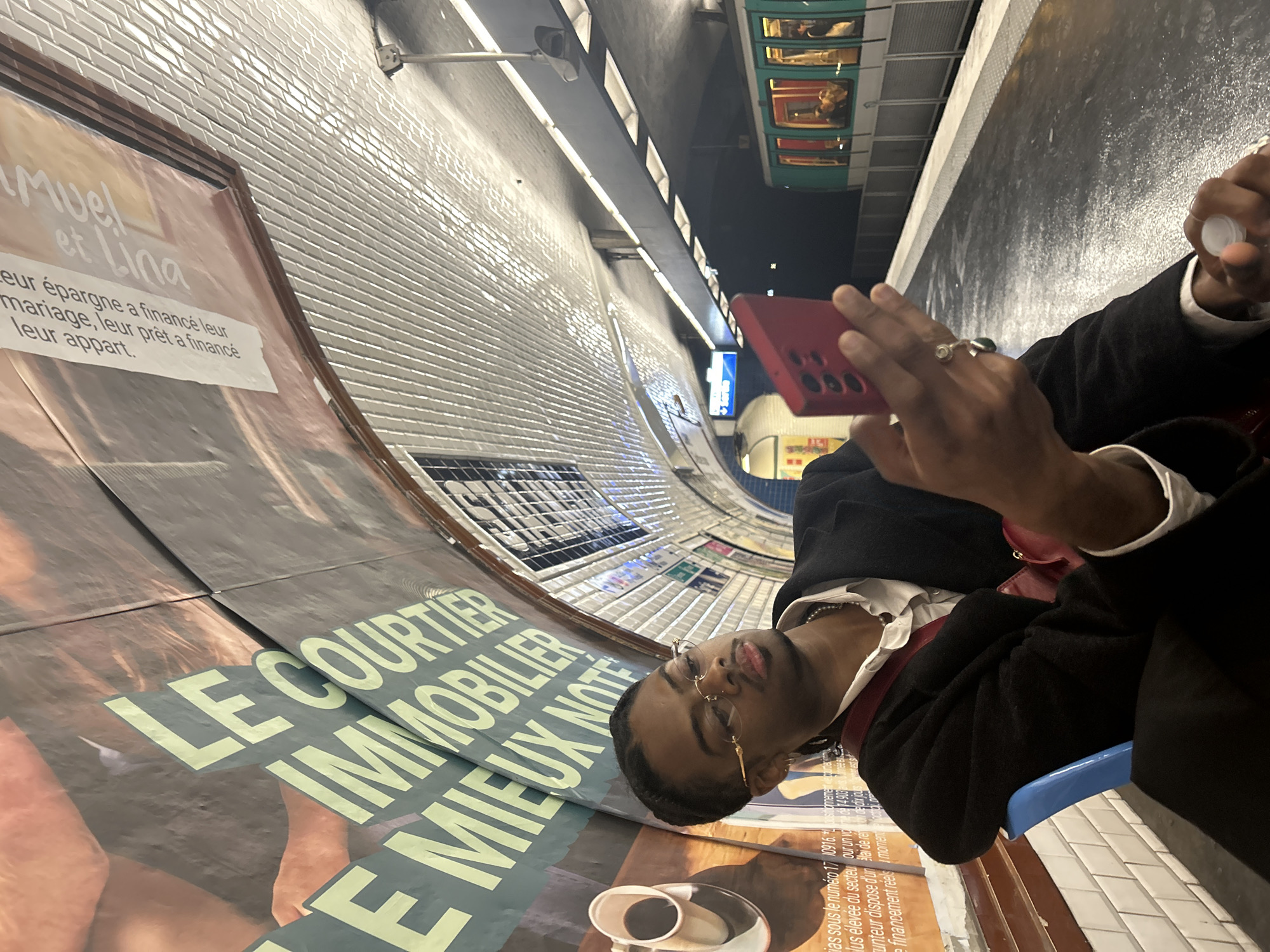 The image shows a man sitting on a blue plastic seat in what appears to be a Paris Metro station, based on the distinctive white tiled curved walls and the presence of a Metro train visible above. The man is wearing a dark blazer over a white shirt with a pearl necklace, and distinctive gold-framed glasses. He is holding a red smartphone in his right hand, and a small white container in his left hand. There's a large advertising poster visible on the wall next to him, reading "LE COURTIER IMMOBILIER E MIEUX NOTE," among other French text. The angle of the photo is somewhat tilted.