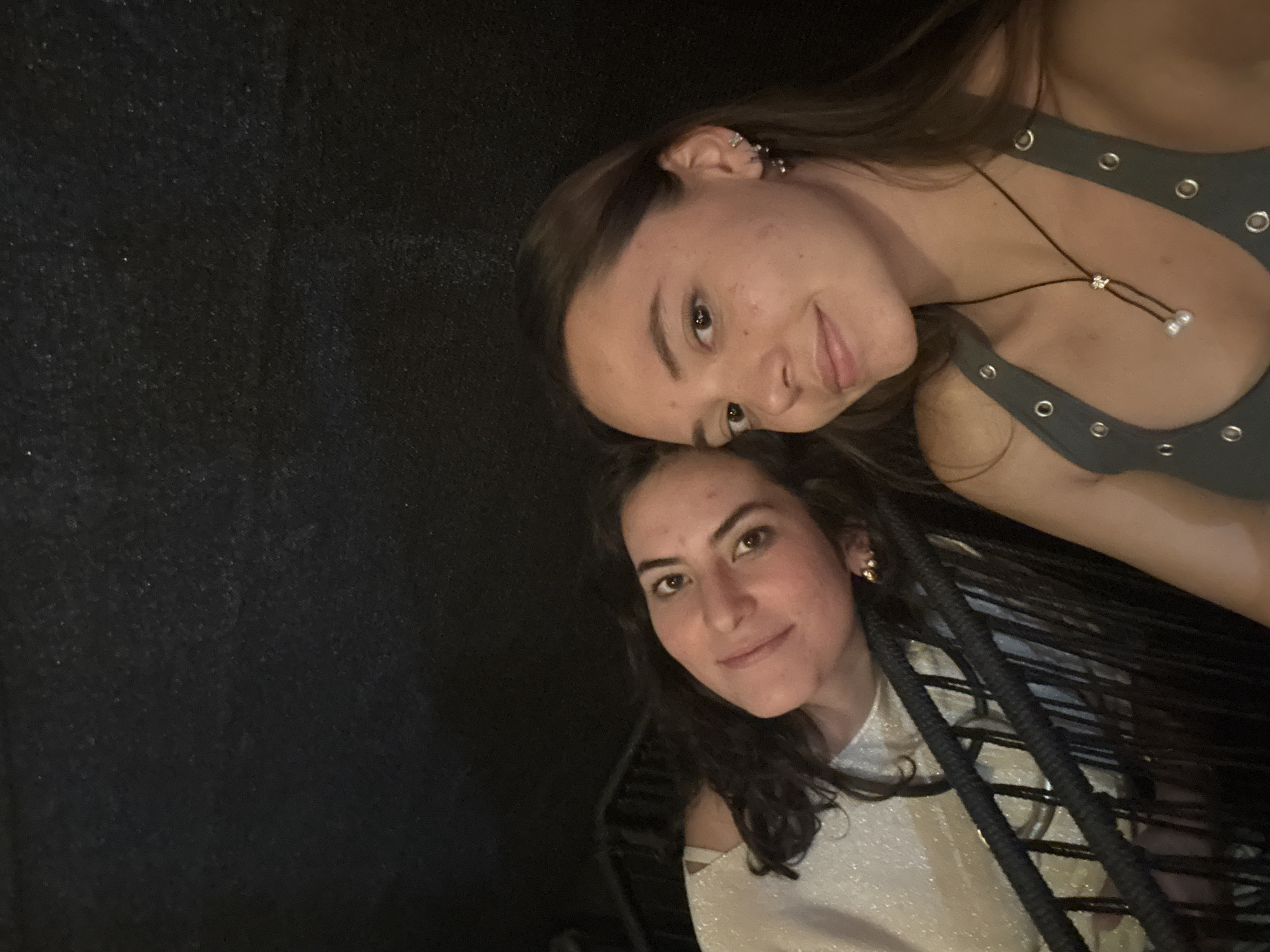 Two young women are posing close together. The first woman, positioned at the top, has shoulder-length brown hair, fair skin, and wears a gray tank top with metallic eyelets. She has multiple ear piercings on her left ear and is wearing a brown cord necklace with decorative accents. The second woman, located below, has shoulder-length dark hair and is wearing a light-colored, shimmering top. The background to the left is black. Braided dark cords partially frame the woman at the bottom right. The location is unclear but is requested as San Bernardino Tlaxcalancingo, Mexico.