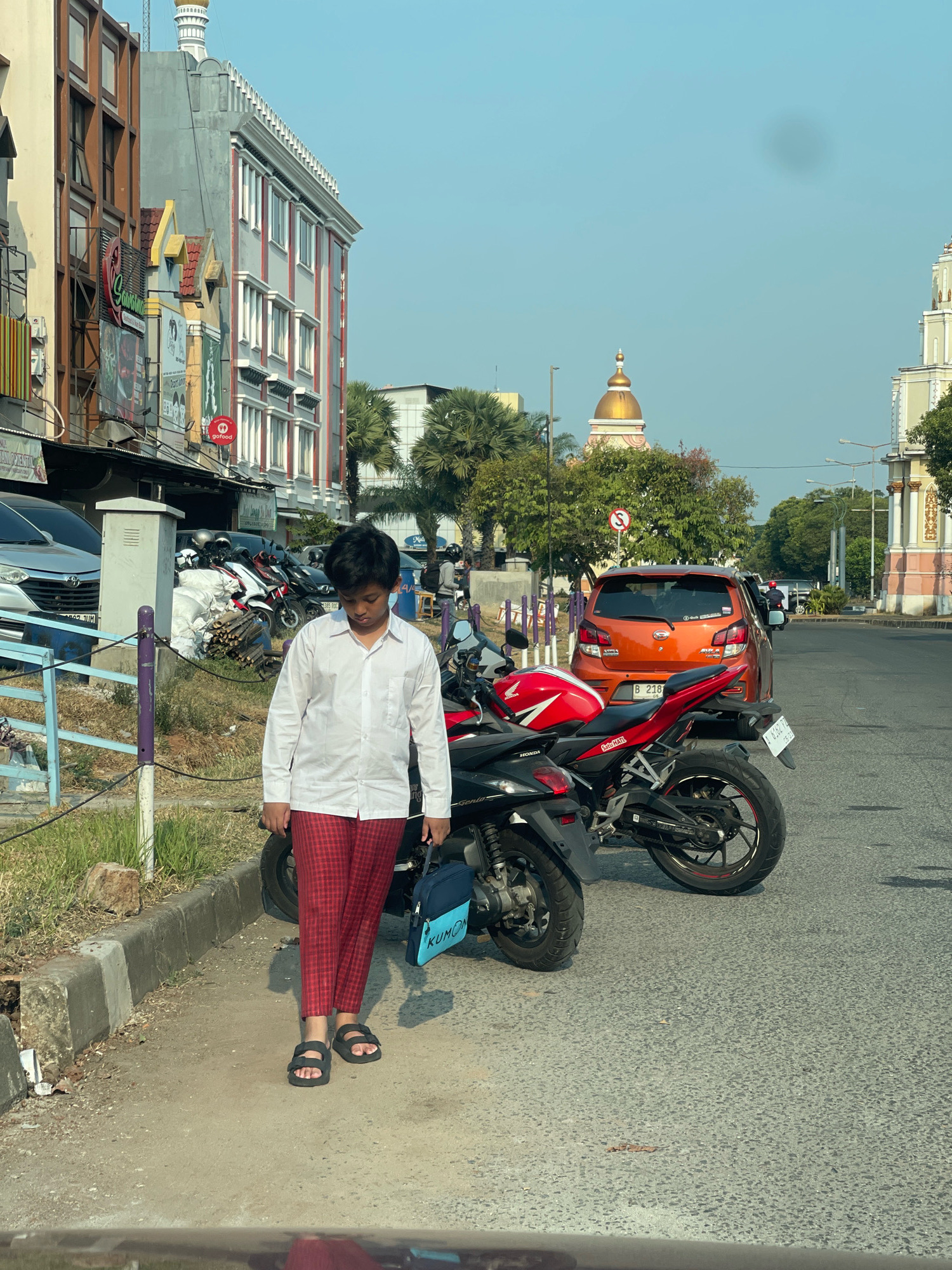 A young boy in a white long-sleeved shirt and red checkered pants walks on the sidewalk next to a street in Pakulonan Barat, Indonesia. He is carrying a blue bag with the word "KUMON" on it. Two motorcycles are parked on the street, and an orange car is driving away. The buildings in the background appear to be shops and offices, with a mosque dome visible in the distance. The sky is clear and blue.