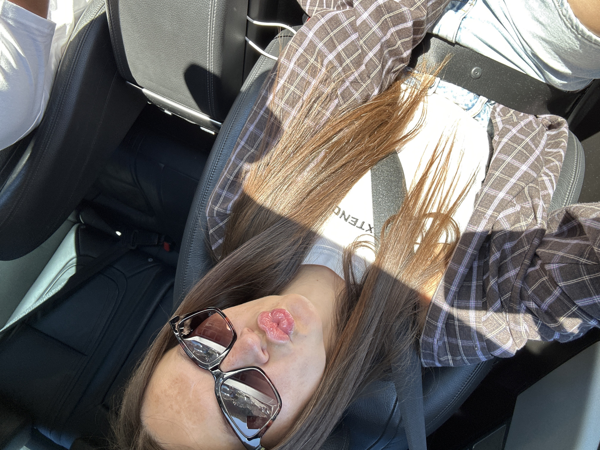 A young woman with long brown hair and sunglasses is taking a selfie in the passenger seat of a car. She is wearing a plaid shirt over a white t-shirt and denim shorts. Her lips are pursed in a kiss. The interior of the car is visible, with black leather seats and a seatbelt. The lighting suggests it is daytime, with sunlight casting shadows. The location is identified as Donji Pobori, Montenegro.
