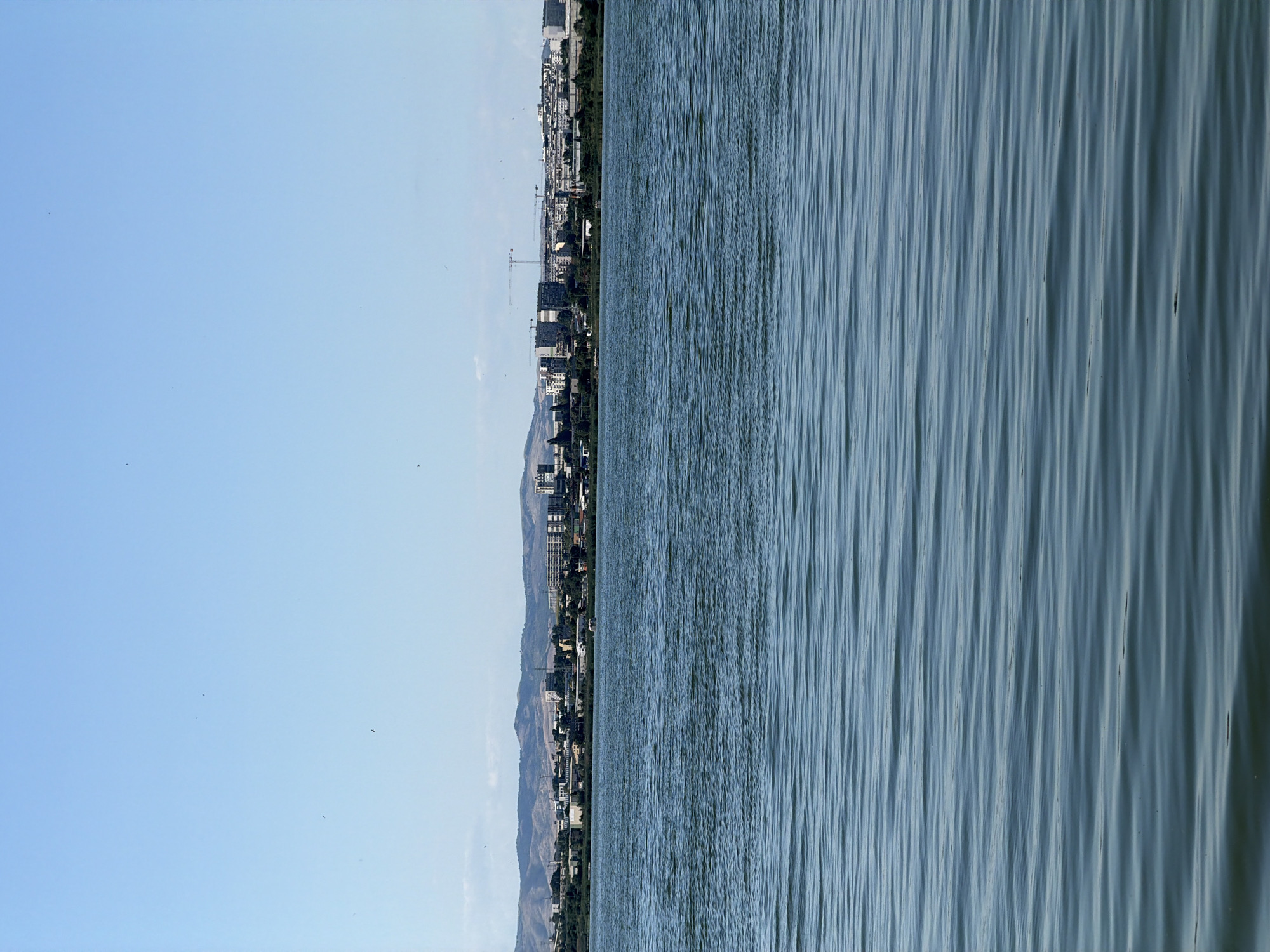 The image captures a scenic view of a city skyline across a body of water, likely a lake or bay, in Burgas, Bulgaria. The composition is split vertically, with the left side dominated by a clear, pale blue sky dotted with a few distant birds and some faint clouds on the far right. The right side of the image is filled with the textured surface of the water, characterized by numerous rippling waves that create a sense of movement and depth.
Along the horizon, separating the sky and water, lies a densely populated urban area. The city is characterized by a mix of residential buildings and taller structures, suggesting a developed cityscape. The presence of several construction cranes indicates ongoing development or urban expansion. Behind the city, rolling hills and mountains are visible, adding a natural backdrop to the urban landscape. The overall scene suggests a daytime setting, with bright, clear weather. The calm, yet dynamic, water surface combined with the distant city and mountains evokes a sense of tranquility and urban life coexisting with nature. No visible text is present in the image.
