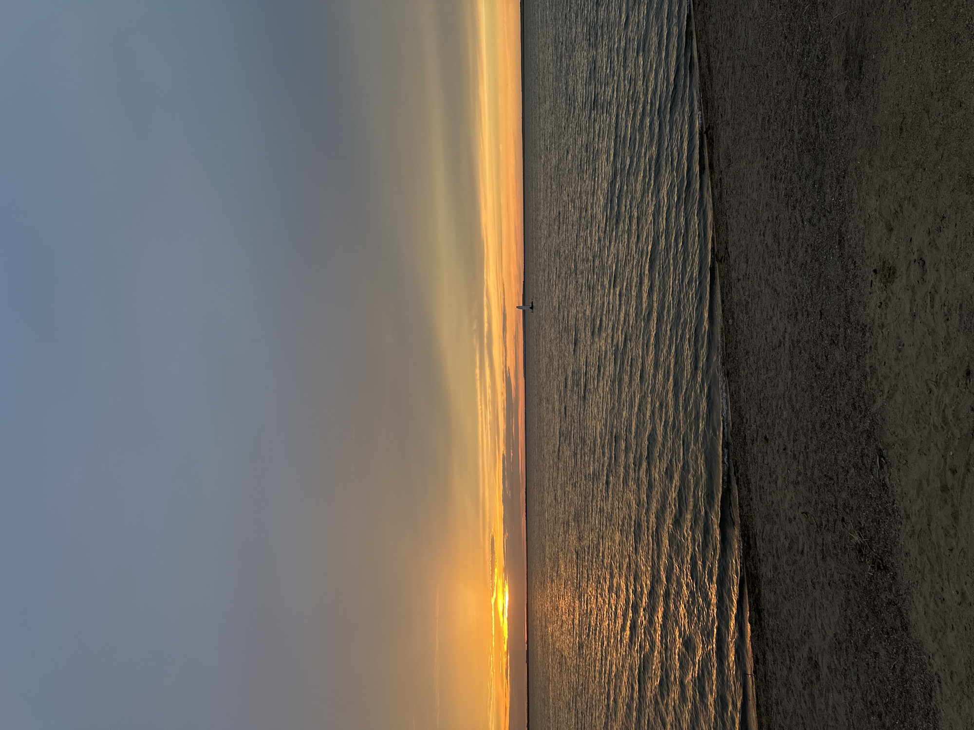A sunset over the ocean creates a gradient of warm colors in the sky, ranging from pale blue to golden yellow and orange. Wispy clouds are present, catching the light. A sailboat is visible in the distance on the water. The water itself is rippled, reflecting the sunset's glow. The foreground shows a sandy beach. The image is taken from a high angle, looking down towards the water and beach. The time of day is sunset, indicated by the warm light and colors in the sky and on the water. The weather appears calm. The scene evokes a sense of tranquility and natural beauty. No text is visible in the image.