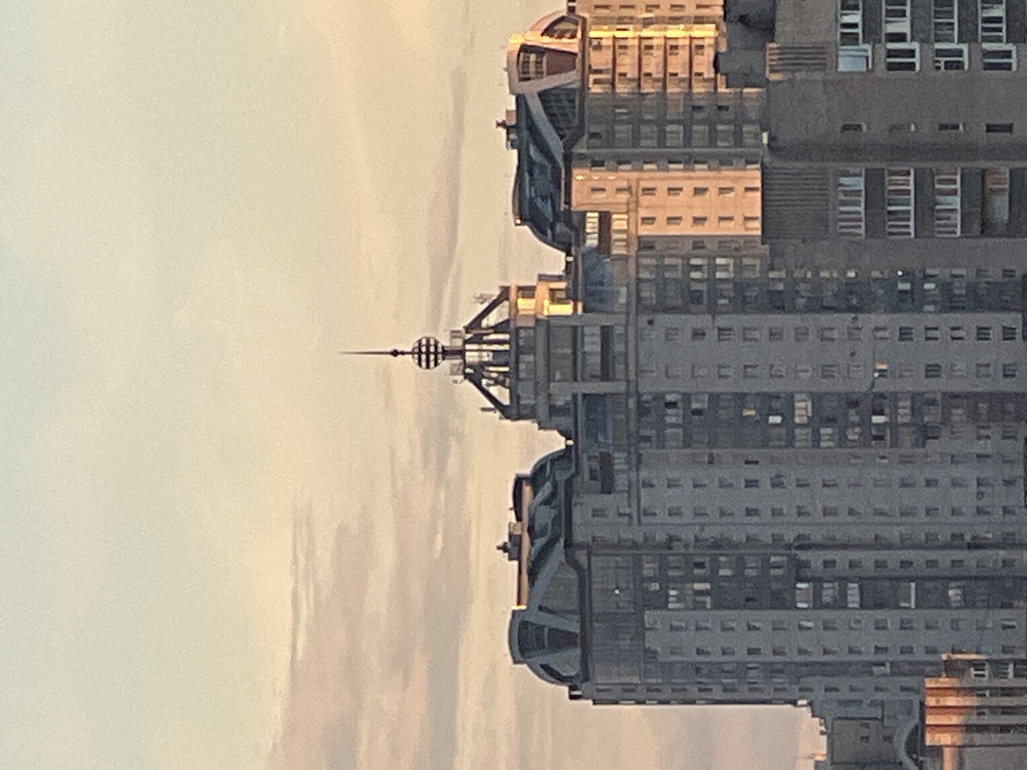 This is a vertical, close-up, eye-level, medium shot of a tall, modern building, likely a skyscraper, bathed in the warm glow of sunset or sunrise. The building is predominantly grey concrete with many windows. The light highlights certain sections of the facade, giving it a warm, golden hue, particularly on the upper levels and a prominent, curved architectural feature.
The building appears to have a complex, multi-tiered structure. One section has a distinctive rounded, almost organic shape. Another section features a metallic spire or antenna with a spherical top, reminiscent of old-fashioned radio antennas or decorative architectural elements. This spire has a series of horizontal bands around its spherical component.
The building's facade is characterized by rows and columns of windows, many of which are rectangular and appear to be uniformly spaced. Some windows are dark, reflecting the sky, while others are lighter, possibly illuminated from within or reflecting the warm exterior light.
The sky in the background is a pale, creamy yellow, indicating either early morning or late evening. There are subtle cloud formations or atmospheric haziness that contribute to the soft lighting. The overall impression is one of urban architecture against a serene, natural light. There is no visible text in the image. The scene is devoid of people, focusing solely on the architectural subject and its interaction with the light.