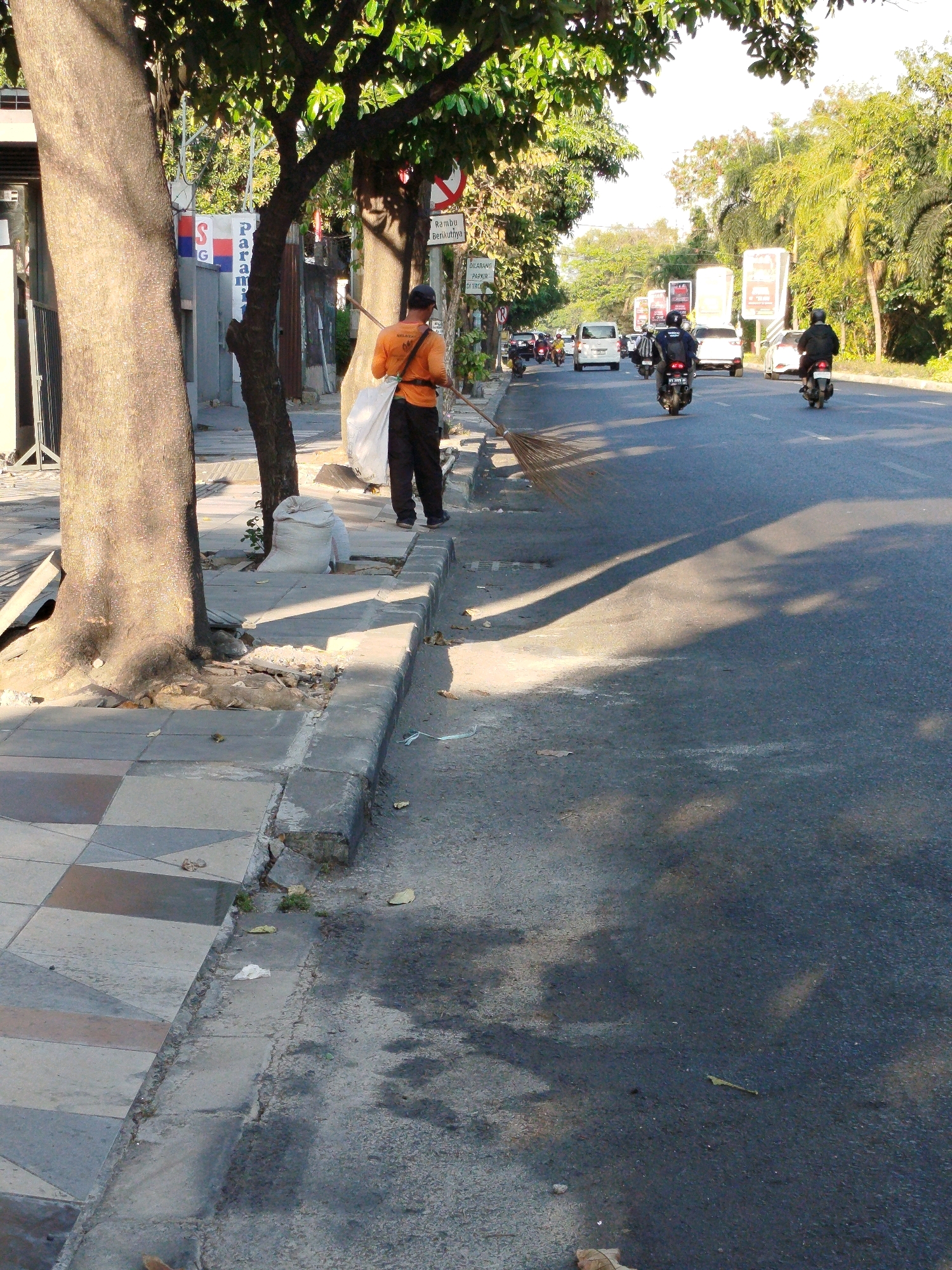 A street cleaner in an orange uniform with black pants and a dark cap sweeps the sidewalk and road with a broom. He has a large white bag next to him. He is standing on the right side of the sidewalk, facing the road. In the background, a street with several cars and motorcycles is visible. There are trees lining the street, and some buildings can be seen in the distance. The scene appears to be in Surabaya, Indonesia, given the presence of the Rambu Lalu Lintas (traffic signs) visible in the background. The sun is shining, casting shadows on the road. The sidewalk is made of concrete and tiles. The road is asphalt. There is some debris on the sidewalk.
