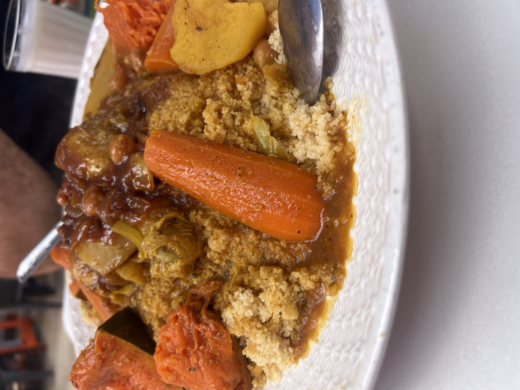 This close-up image presents a traditional North African meal, likely couscous with a vegetable and stew, served on a white plate with a textured rim. The dish features a generous portion of fluffy, yellow-tinted couscous, partially covered by a rich, savory brown stew.
Prominent elements within the stew include several large, tender-looking orange carrots, some appearing whole or halved. Other stewed vegetables are visible, such as chunks of yellow potato or squash, and lighter green-yellow pieces that could be celery or cabbage. The stew itself is thick and saucy, with discernible pieces of cooked onion and other ingredients.
A silver utensil, possibly a spoon or fork, is resting in the food on the right side of the plate, suggesting the meal is in progress or about to be enjoyed. The plate is positioned diagonally, with its patterned rim visible.
In the blurred background on the left, part of a person's arm or clothing and a clear glass or cup can be faintly seen, indicating an indoor dining environment. The right side of the background shows a plain, light-colored surface, likely a table. The characteristics of the meal are consistent with Moroccan cuisine, aligning with the provided context of Casablanca, Morocco. No text is visible in the image.