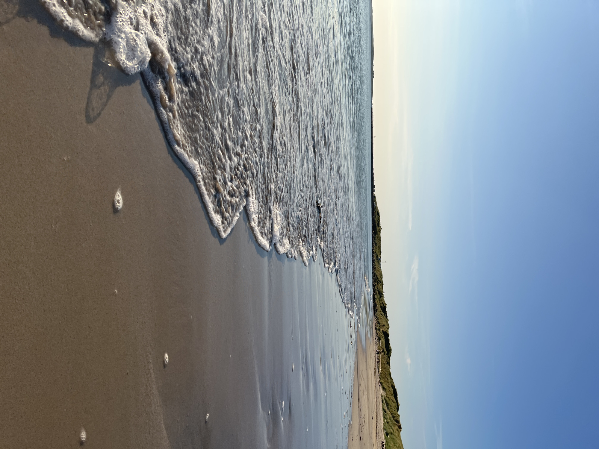 An eye-level, medium shot captures a section of the beach at Lunniaghbeg, Ireland, with the sea on the left and the shore on the right. The sky is a clear, light blue, stretching across the top of the frame. In the distance, a low, green, grassy dune system runs along the coastline, with a few indistinct figures visible walking on the sand. The sea is calm, with small waves gently lapping the shore. The wet sand reflects the sky, creating a mirror-like surface. The overall atmosphere is tranquil and serene.