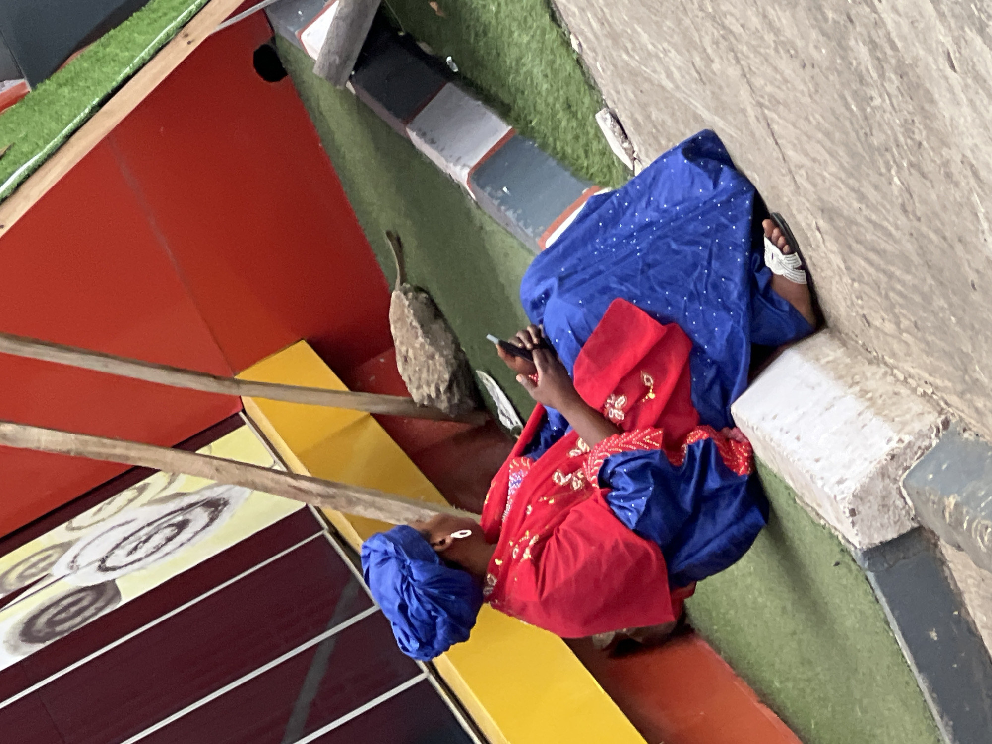 The image depicts a person in Gura Top, Nigeria, wearing a traditional blue and red outfit, seated on a structure with red, yellow, and maroon elements. The person is using a mobile phone. The backdrop includes artificial green turf and concrete or stone elements.