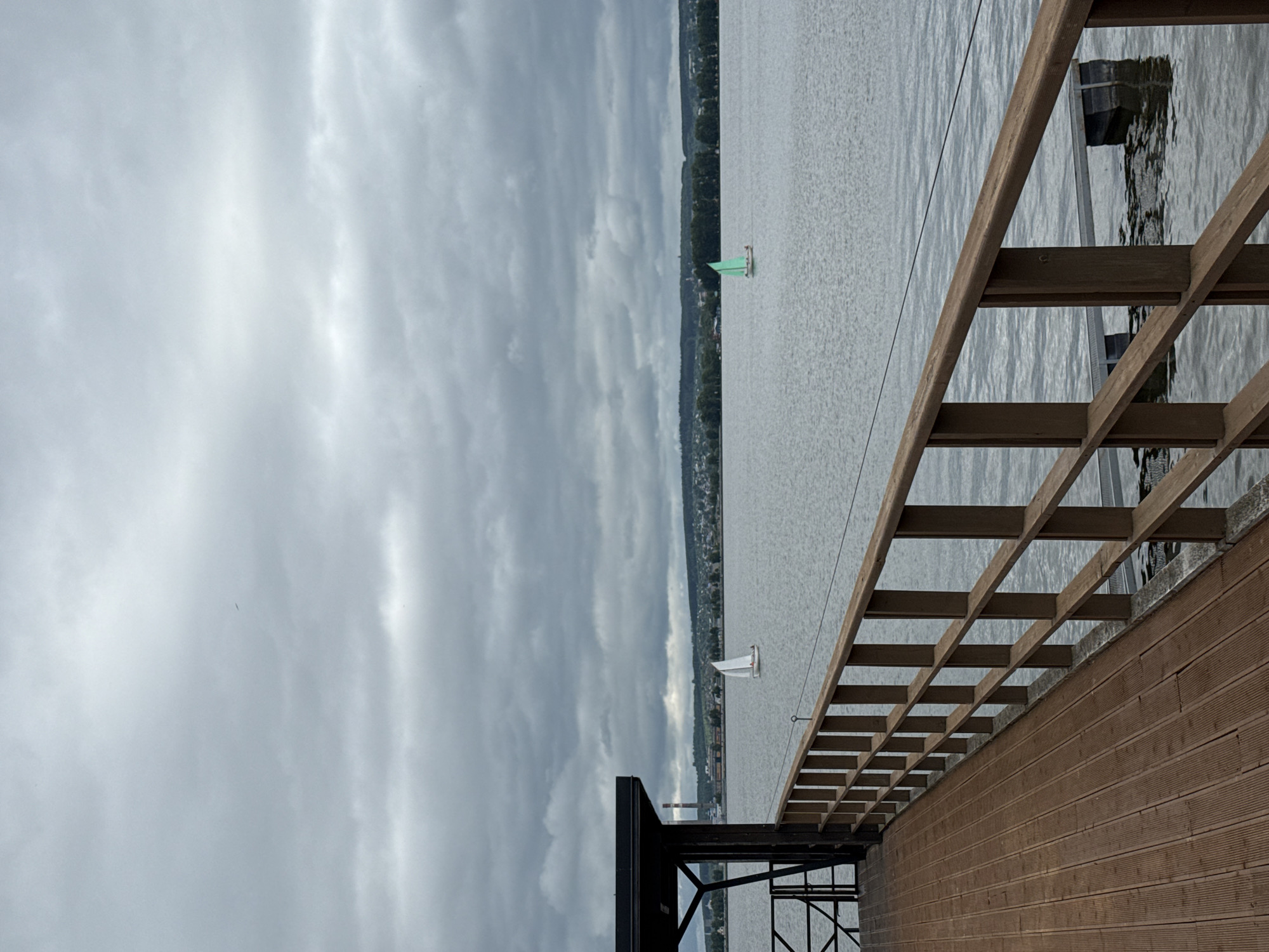 The image captures a wide view of the Votkinsk Reservoir in Votkinsk, Russia, under an overcast sky. In the immediate foreground, a dark brown wooden pier with horizontally slatted planks extends from the lower left, accompanied by a robust, lighter brown wooden railing running along its edge. A dark-roofed structure with black metal supports is partially visible on the far left. Two small sailboats are positioned on the calm, grey water: one with a white sail is situated towards the left-midground, while another with a distinct mint green sail is further right. Each boat appears to contain at least one indistinct individual. The distant shoreline reveals a tree-lined horizon, scattered buildings indicative of a town or city, and a prominent tall, light-colored industrial chimney on the left. The entire upper half of the frame is dominated by a dense, uniform layer of grey stratocumulus clouds, with a single minute dark speck visible in the upper right. A dark floating platform is partially submerged beyond the railing in the lower right water.