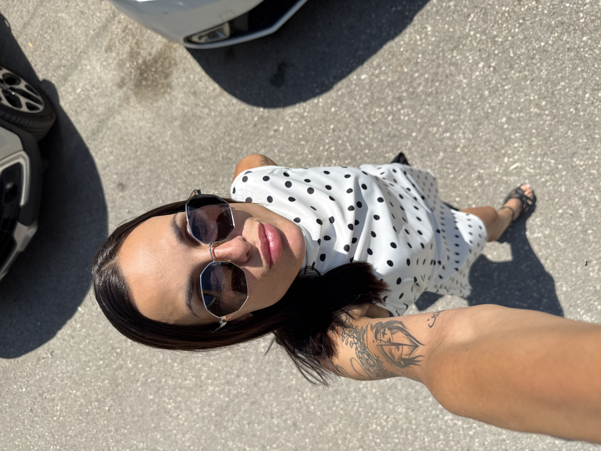 The image features a woman standing on an asphalt surface, likely a parking lot, with two cars visible to her left and top. She is wearing a white dress with black polka dots and black sandals. The woman is looking up towards the camera, wearing sunglasses and her lips are slightly parted. Her hair is dark brown and falls around her shoulders. She has tattoos on her right arm. The scene is illuminated by bright sunlight, suggesting it is daytime, and the shadows cast by the woman and the cars indicate a sunny weather condition. The setting appears to be outdoors, possibly in a town or city, given the presence of cars and paved ground. The overall mood of the image seems casual and relaxed. No visible text is present in the image. The location context provided is Pleven, Bulgaria.