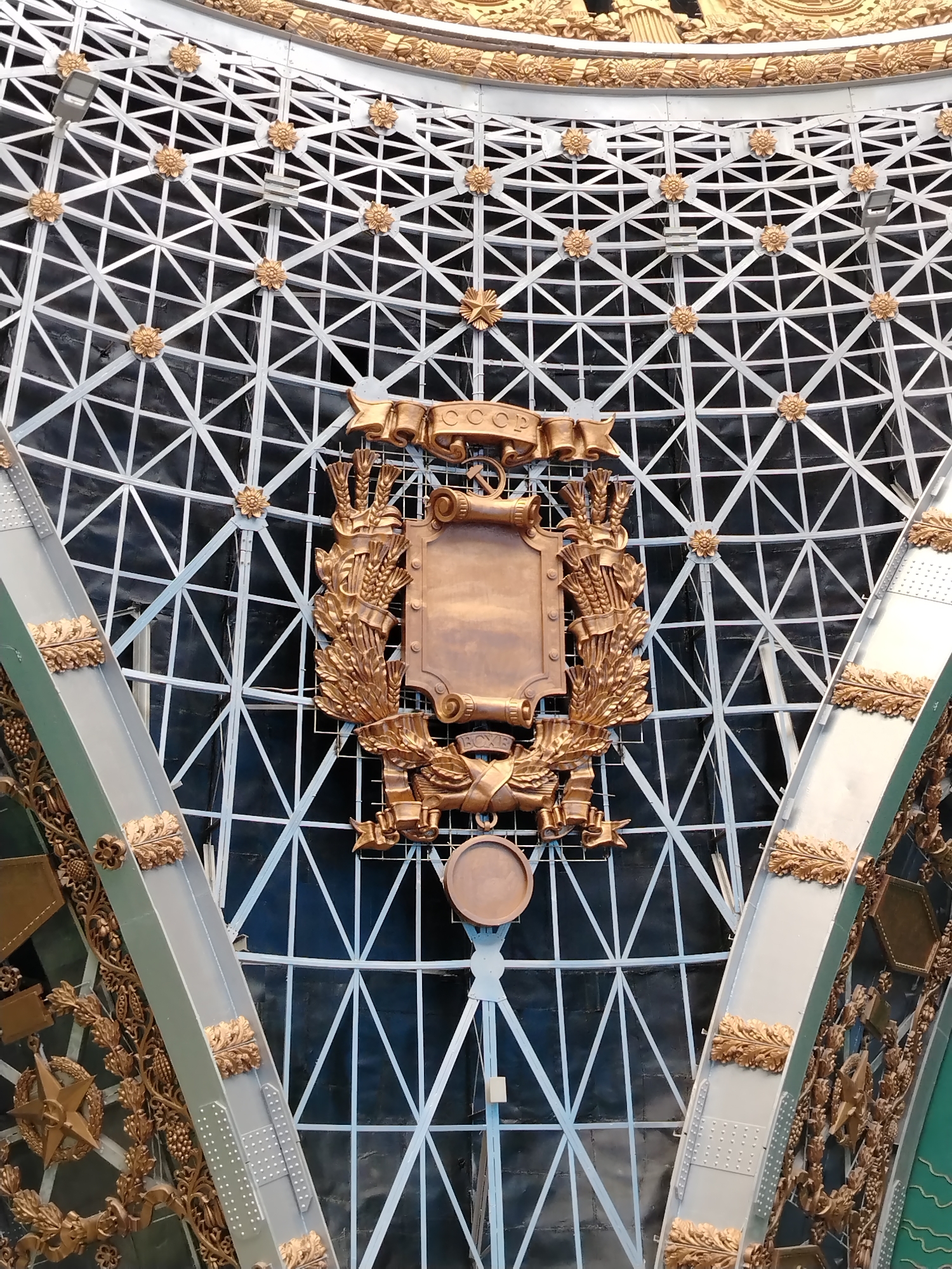 This image depicts a close-up view of the golden emblem of the Soviet Union, displayed on the facade of the All-Russia Exhibition Centre (VDNKh) in Moscow, Russia. The emblem, a shield surrounded by wheat stalks and featuring a hammer and sickle, is prominently featured against a backdrop of a white, lattice-like metal structure. Decorative golden elements, including ribbons and stars, are also visible as part of the overall facade design. The scene appears to be an outdoor setting, with natural light illuminating the metallic surfaces of the emblem and surrounding architecture.