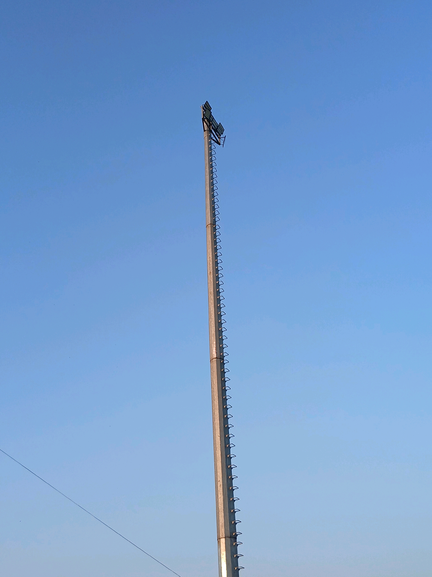 A tall concrete pole with a ladder attached to its side stands against a clear blue sky. At the top of the pole, there are two floodlights mounted on a metal frame. The pole is located outdoors in a bright, open area, likely a field or a sports complex, given the presence of the floodlights. The sky is cloudless, indicating fair weather. A thin wire runs diagonally across the lower left portion of the image. The image does not contain any people.