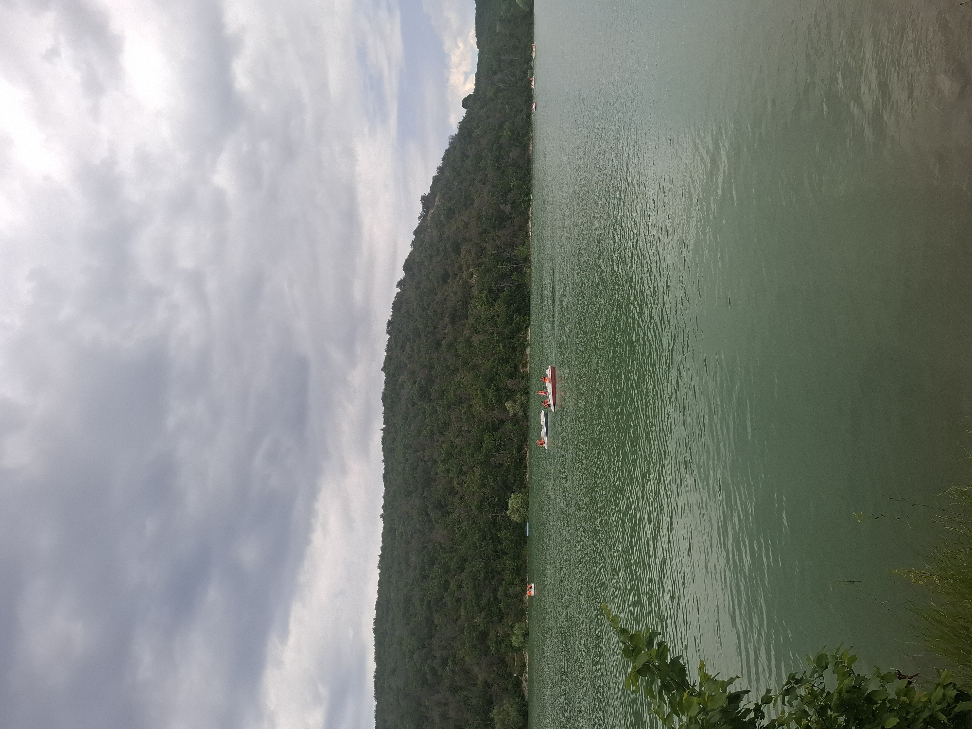 The image depicts a wide view of a lake in the city of Sukko, Russia. The lake water is a murky green color, with small ripples across its surface. In the distance, a densely forested hill forms the backdrop to the scene. Several small paddle boats, some with people wearing orange life vests, are scattered across the lake. The sky above is overcast with gray clouds, suggesting a potentially cloudy or rainy day. In the foreground, the edge of the lake shows some green vegetation and grass.