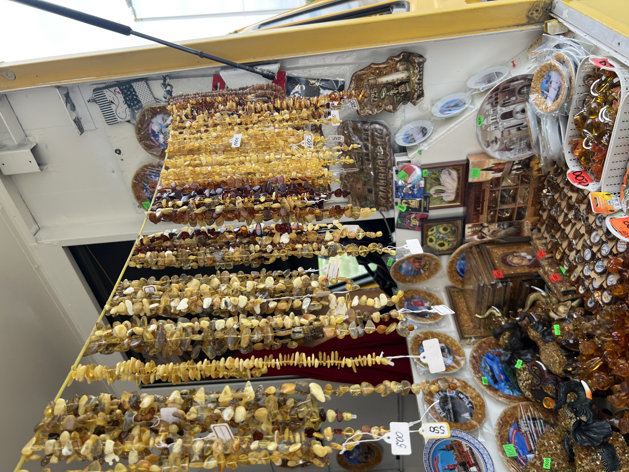 The image depicts a street vendor stall in Baltiysk, Russia, specializing in amber products. Strings of various amber beads, some polished and others rough, hang from a yellow rod, catching the light. These range in color from pale yellow to deep brown. Below the hanging beads, a variety of amber-encrusted items are displayed, including elephant figurines, decorative plates with scenic depictions of Russian landmarks such as St. Basil's Cathedral, and small framed artworks featuring swans and sunflowers. Several price tags are visible, with denominations like "500," "600," "800," "1500," and "200." A yellow truck or van is partially visible on the right side of the frame, suggesting the stall might be mobile. The background shows a white wall and a glimpse of an outdoor environment, likely a street or market area. There are no visible people actively interacting with the stall at this moment.
