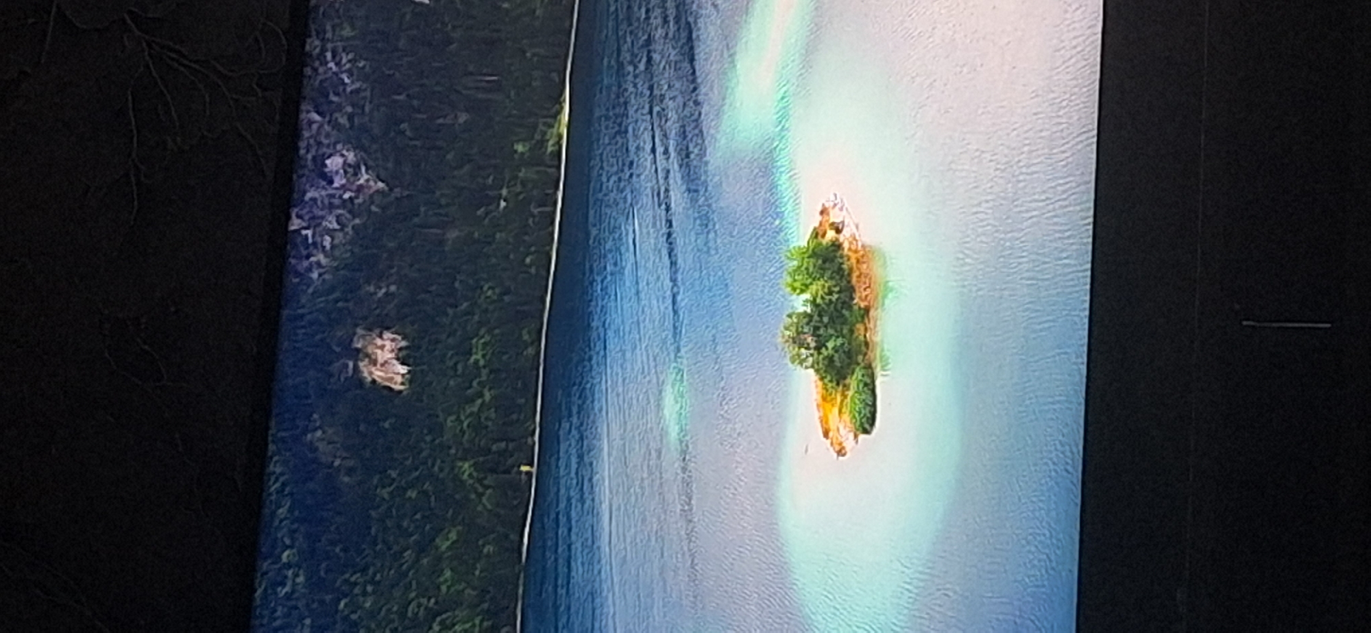 The image shows a picturesque landscape with a small, verdant island in the center of a calm body of water. The island is covered with lush green trees and some patches of earth. The water surrounding the island is a striking turquoise color, with ripples creating a shimmering effect. In the background, a dense forest covers a steep hillside, with a few rocky outcrops visible. A thin, white line, possibly a path or a shore, runs along the base of the hillside. The overall scene conveys a sense of tranquility and natural beauty. The provided information about the city of Bila Tserkva, Ukraine, does not directly correlate with the visual content of the image, as the scene depicts a natural landscape that is not specifically identifiable as being located within that city without further context.