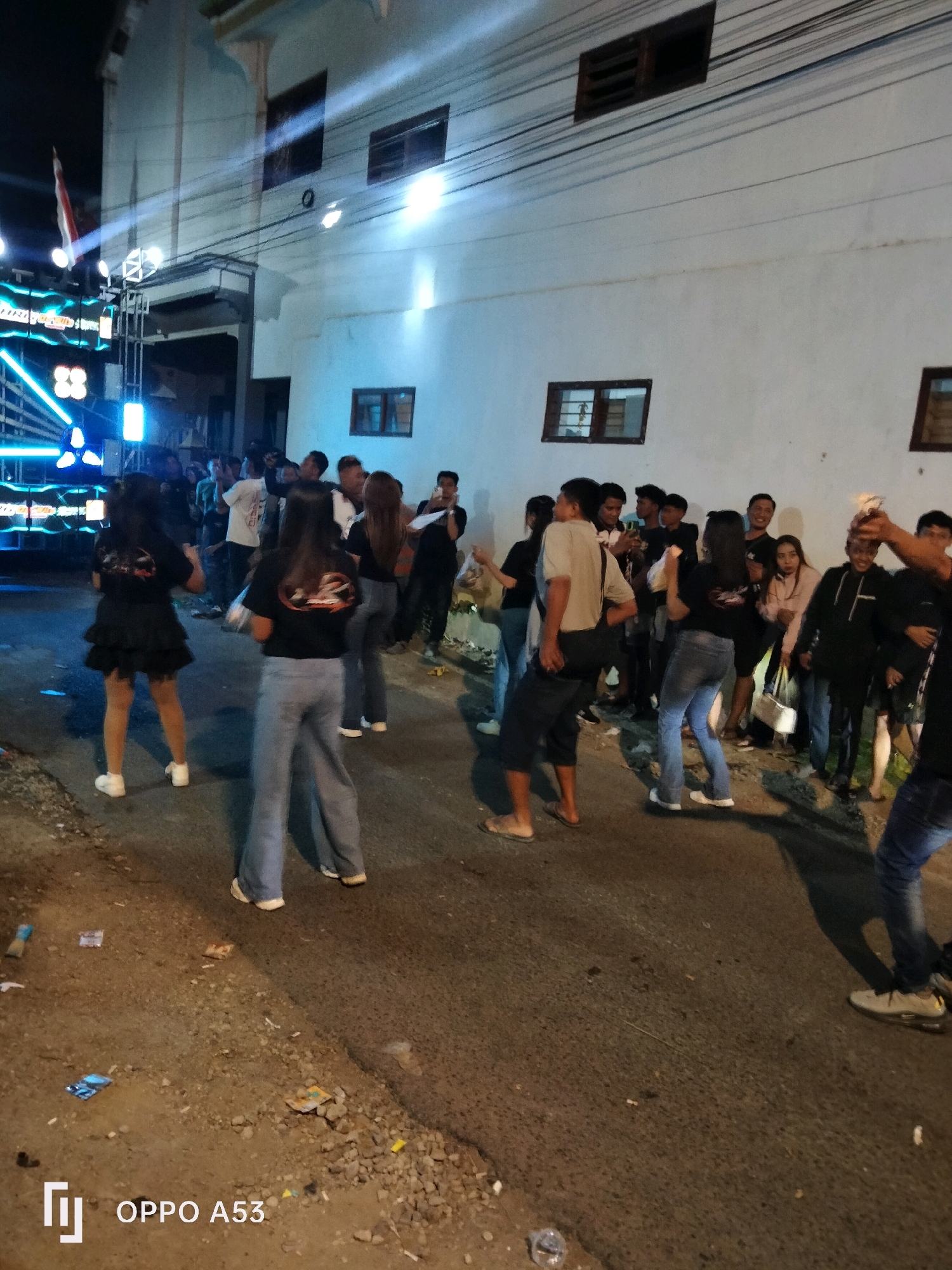 The image depicts an outdoor nighttime gathering in Sukorame, Indonesia, possibly a street party or event, as indicated by the presence of a sound system with bright blue lights on the left and a crowd of people. The scene is illuminated by artificial lighting, including spotlights that cast beams across the white building facade in the background. A white building with multiple windows and what appears to be a covered entryway occupies the upper portion of the frame. A flag, partially visible on a flagpole near the sound system, is a prominent detail in the upper left. The ground is paved with asphalt and scattered with debris, including paper and plastic waste. Several individuals are seen dancing and interacting, with some holding up their hands as if in celebration or holding objects that might be sparklers. Two women in the foreground are wearing black t-shirts with a logo and appear to be dancing. The overall atmosphere suggests a lively social event.