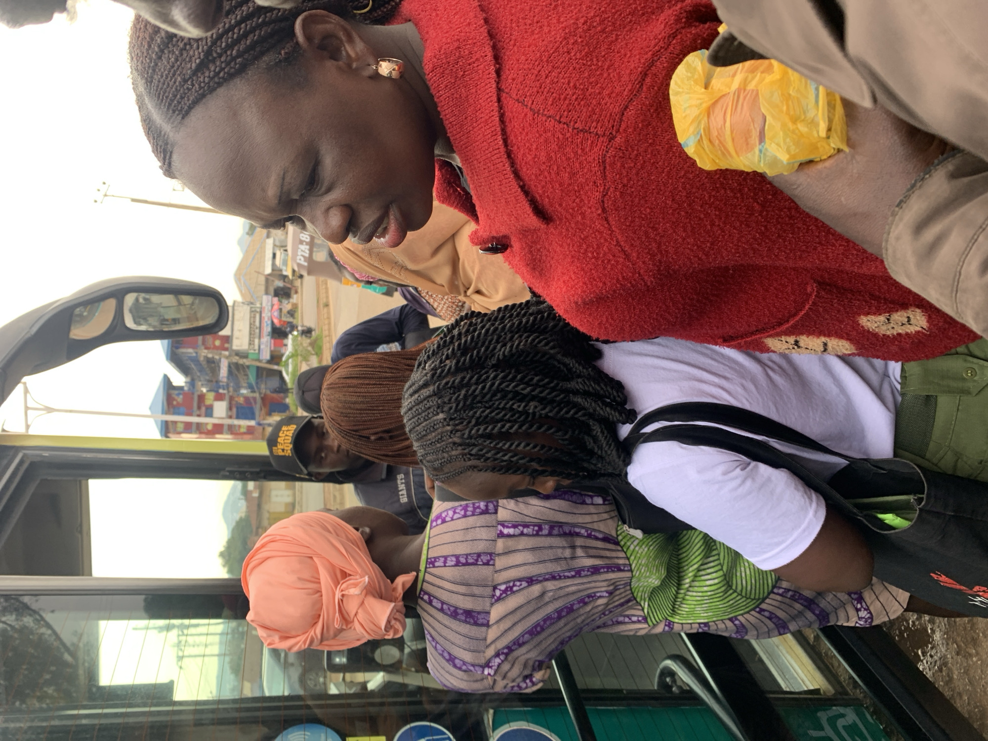 In Rafin Par, Nigeria, a group of individuals, including women and a man, are gathered near the entrance of a bus. The scene is captured in daylight, with a slightly overcast sky. The bus has a glass door and a side mirror, reflecting the surrounding environment, which includes buildings and a road with vehicles.
One woman in the foreground wears a red, textured jacket and has dark skin and braided hair. She is looking to the left, with her mouth slightly open. She holds a crumpled yellow plastic bag in her right hand. To her left, a woman with dark skin and braided hair wears a white t-shirt and green pants, carrying a black tote bag. She is looking down.
Behind her, a woman in a pink headwrap and a patterned dress with purple and white stripes is visible. She faces away from the camera. Another woman with braided brown hair is partially visible behind the woman in the red jacket. In the background, a man wearing a black cap with "GIANTS" written on it is visible. The environment shows a bustling urban setting with multi-story buildings, some with colorful facades, and a paved road. Power lines and streetlights are also present. The ground appears to be damp, suggesting recent rain.