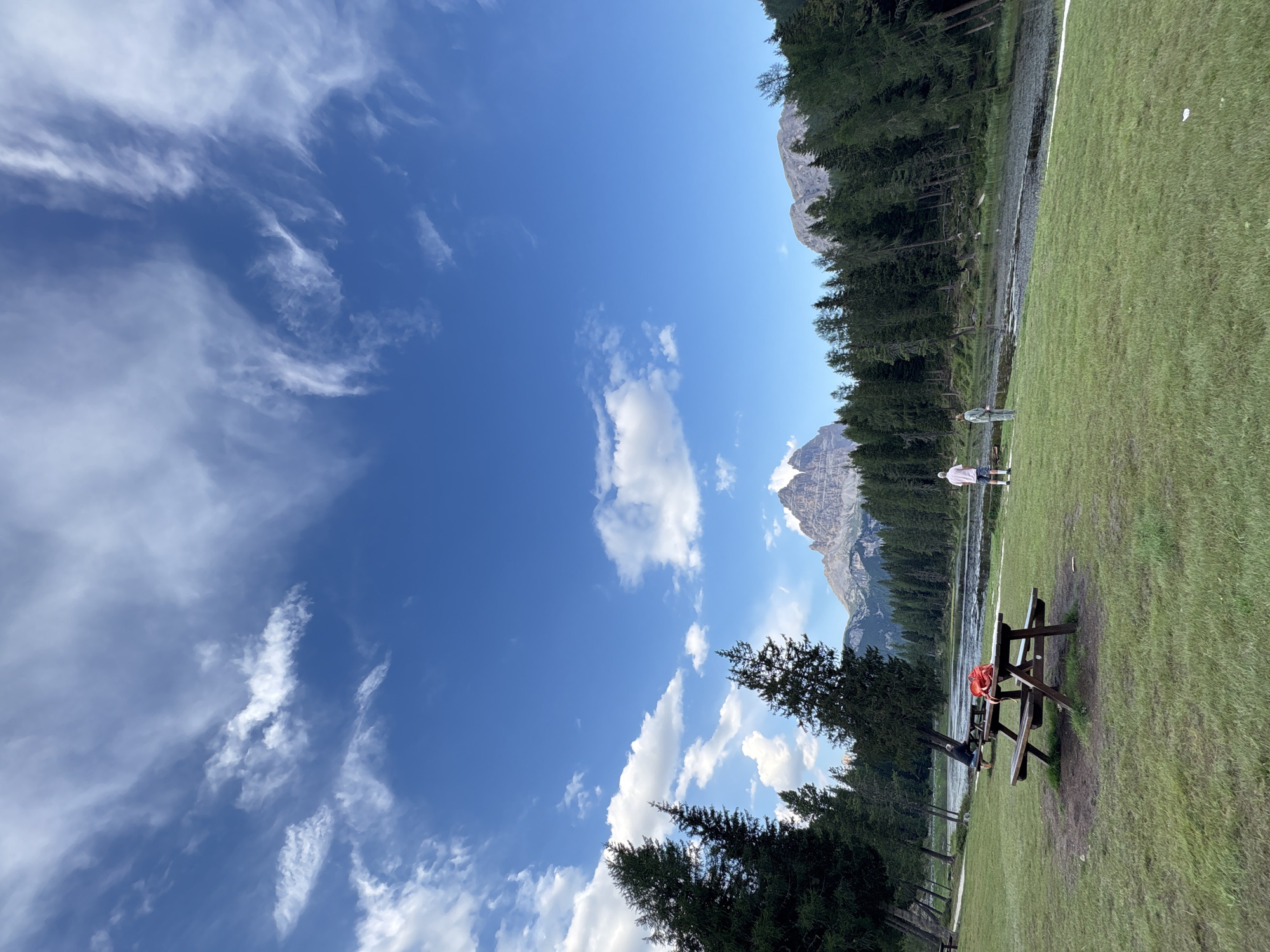 The image depicts a serene landscape in Auronzo di Cadore, Italy, likely near Lago di Misurina. The foreground is a lush green grassy field with a wooden picnic table in the center, upon which rests a red bag. To the right of the picnic table, two individuals are standing in the field, facing the lake. The middle ground features a body of water, possibly a lake, bordered by a dense forest of tall evergreen trees. In the background, imposing mountains with rugged peaks rise majestically against a vibrant blue sky filled with wispy white clouds. The overall scene conveys a sense of tranquility and natural beauty.
