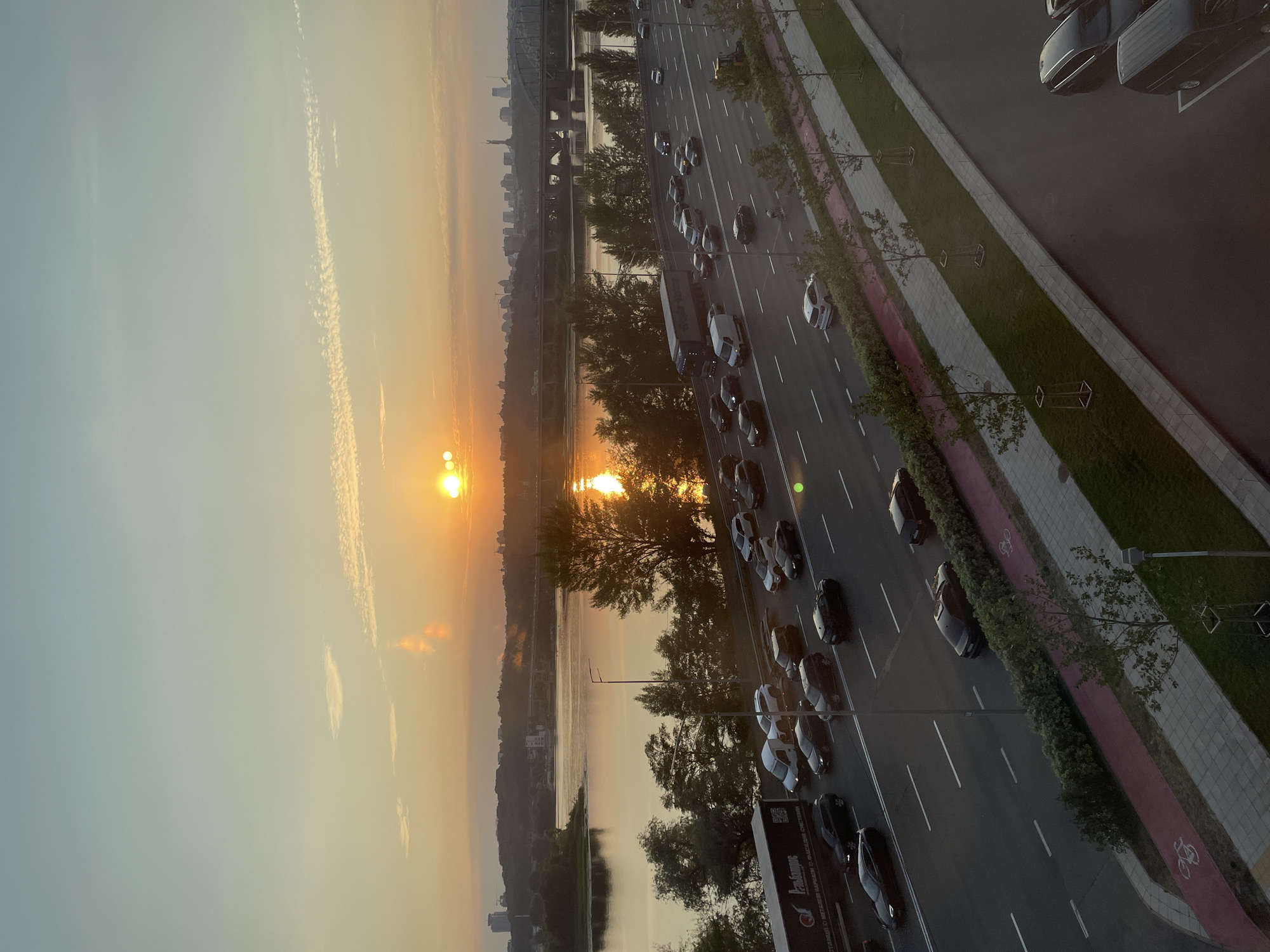 The image captures a wide-angle view of a multi-lane highway and a body of water during sunset in an urban environment. The sun, a bright orb, is positioned in the upper right quadrant of the sky, casting a warm golden glow that reflects off the water and illuminates the clouds. The sky transitions from a pale blue at the top to shades of orange and yellow near the horizon.
On the highway, a steady stream of vehicles, including cars and a large truck, are visible traveling in both directions. The traffic appears to be moving, though some congestion is noticeable. The highway is bordered by trees and a pedestrian/bicycle path. Across the water, a cityscape is visible with buildings and bridges silhouetted against the colorful sky. The water itself is calm, reflecting the warm tones of the sunset. The overall scene suggests a bustling city at the end of the day.