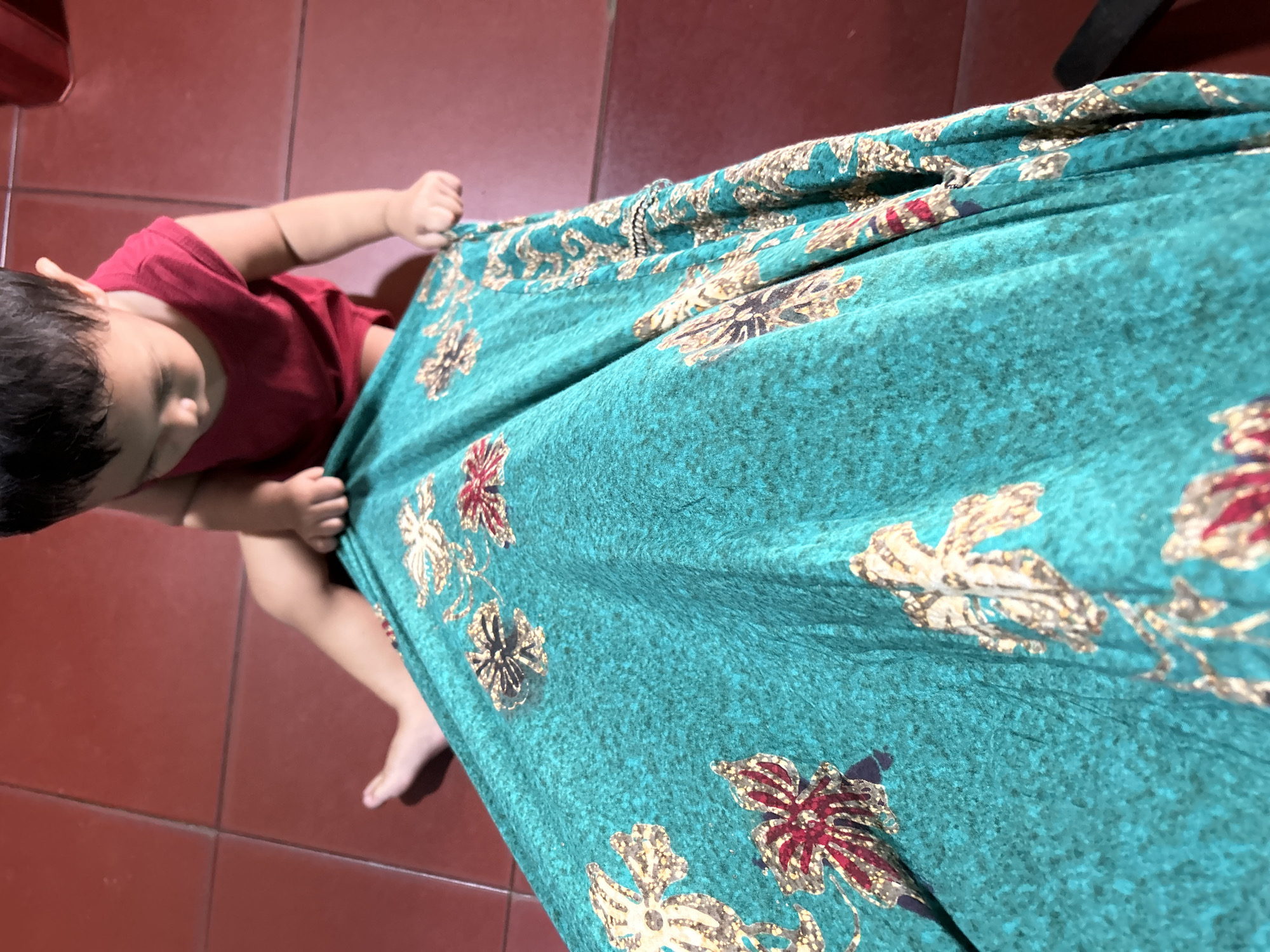 An overhead, slightly angled shot shows a young child in a red sleeveless shirt and a teal fabric with a gold and red floral pattern. The child is seated on a red tiled floor, leaning forward and holding onto the fabric, which covers the lower portion of the frame. The child's bare feet are visible. The tiled floor extends throughout the visible area. The setting appears to be indoors, possibly a home, in Depok, Indonesia.