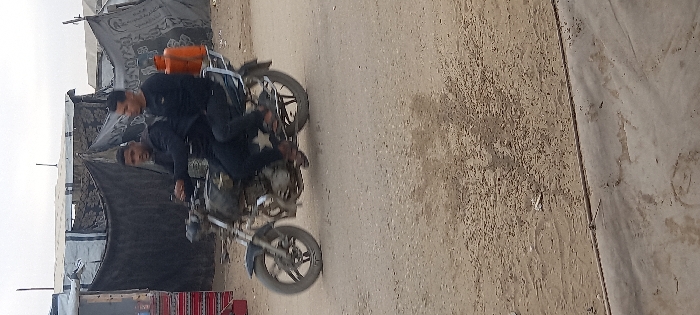 Two adult males are observed riding a motorcycle on an unpaved road in Khan Yunis, Palestinian Territory. The driver, wearing dark attire and light-colored sandals, is positioned forward with hands on the handlebars. The passenger, also in dark attire and sandals, sits behind the driver. An orange gas cylinder is secured on the motorcycle's rear rack. The motorcycle is dark-colored and shows motion blur on its wheels, indicating movement. In the background, multiple makeshift shelters constructed from tarpaulins and dark fabric are visible, suggesting a temporary settlement. To the far left, a small stall with displayed items and a red and white patterned fabric is present. The sky is overcast. The foreground consists of a lighter, possibly concrete, surface transitioning to the dusty, light brown unpaved road.