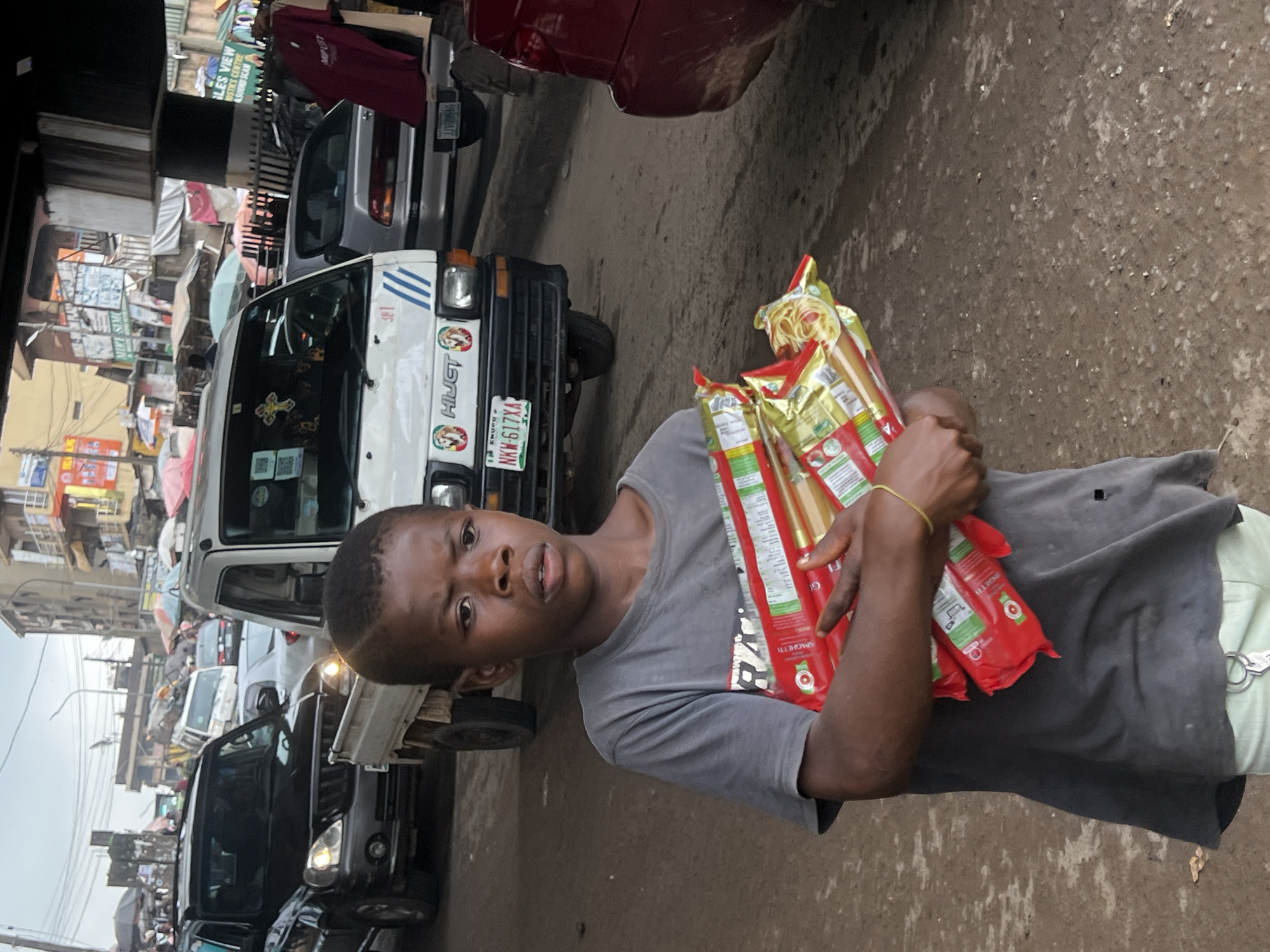 A young boy stands in a busy street in Nkpor, Nigeria, holding multiple packages of spaghetti. The boy, with dark skin and short black hair, wears a gray t-shirt and light-colored pants. He is looking directly at the camera with a neutral expression. He is holding four red packages of spaghetti, with one package opened to reveal the noodles inside.
The background shows a bustling street scene with several vehicles, including a white HiJET truck and a black SUV, indicating a traffic jam. The road surface is dirt and asphalt, with some scattered debris. In the background, buildings with shops and signs are visible, typical of a commercial area in Nigeria. Overhead, a structure, possibly a bridge or overpass, casts a shadow over the scene. The overall environment suggests a lively and crowded urban setting.