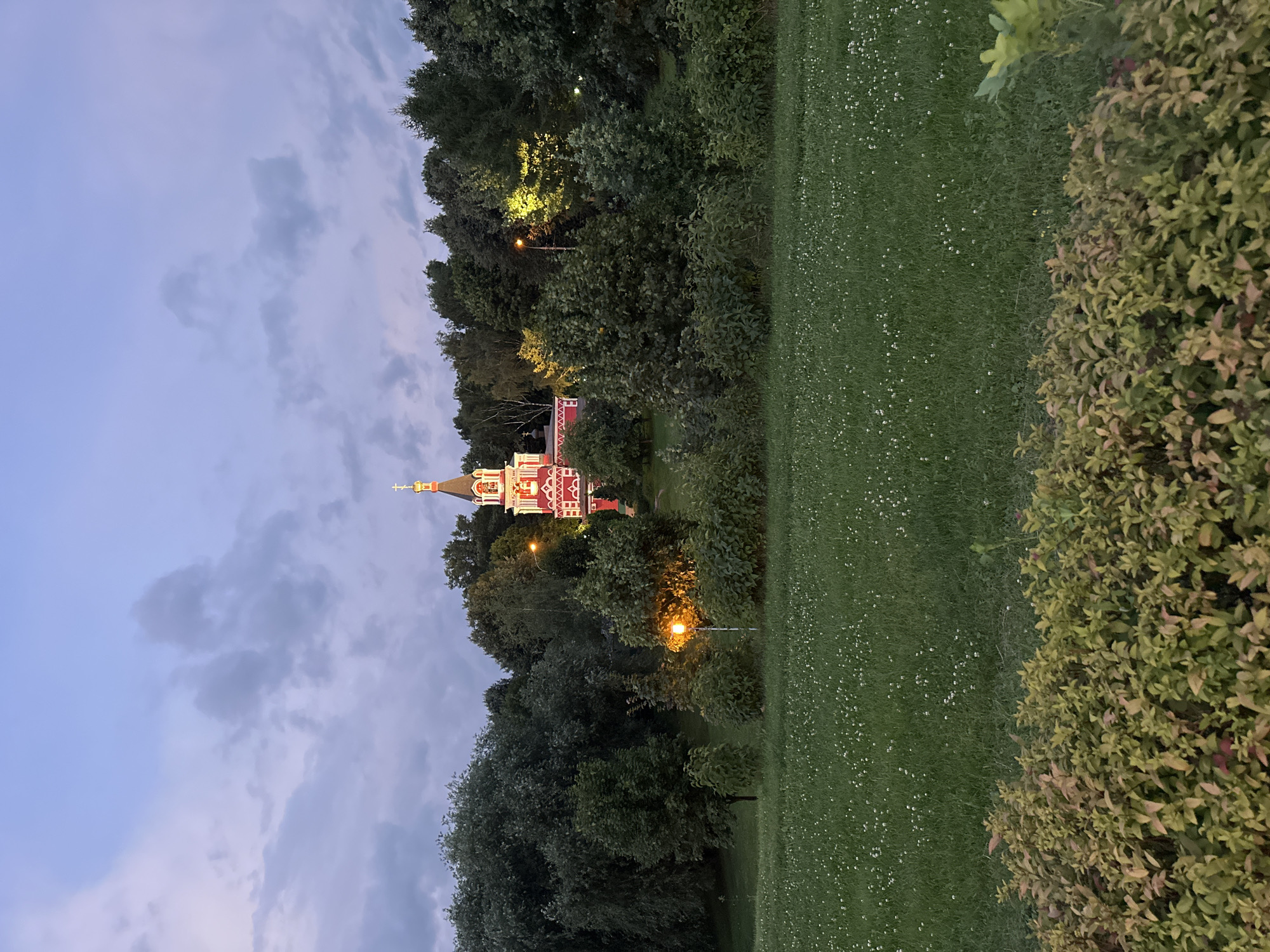A photograph captures an Orthodox church, illuminated by internal and external lighting, standing atop a hill covered in trees. The church is red with white trim and features a golden cross atop its spire. A lamppost in front of the church emits a warm yellow light. The foreground of the image displays a lawn dotted with white clover flowers, bordered by a shrubbery with variegated green and reddish leaves. The sky above is a dusky blue with scattered clouds. This scene is located in Kolomenskoye Park, Moscow, Russia. There are no people or animals visible in the image.