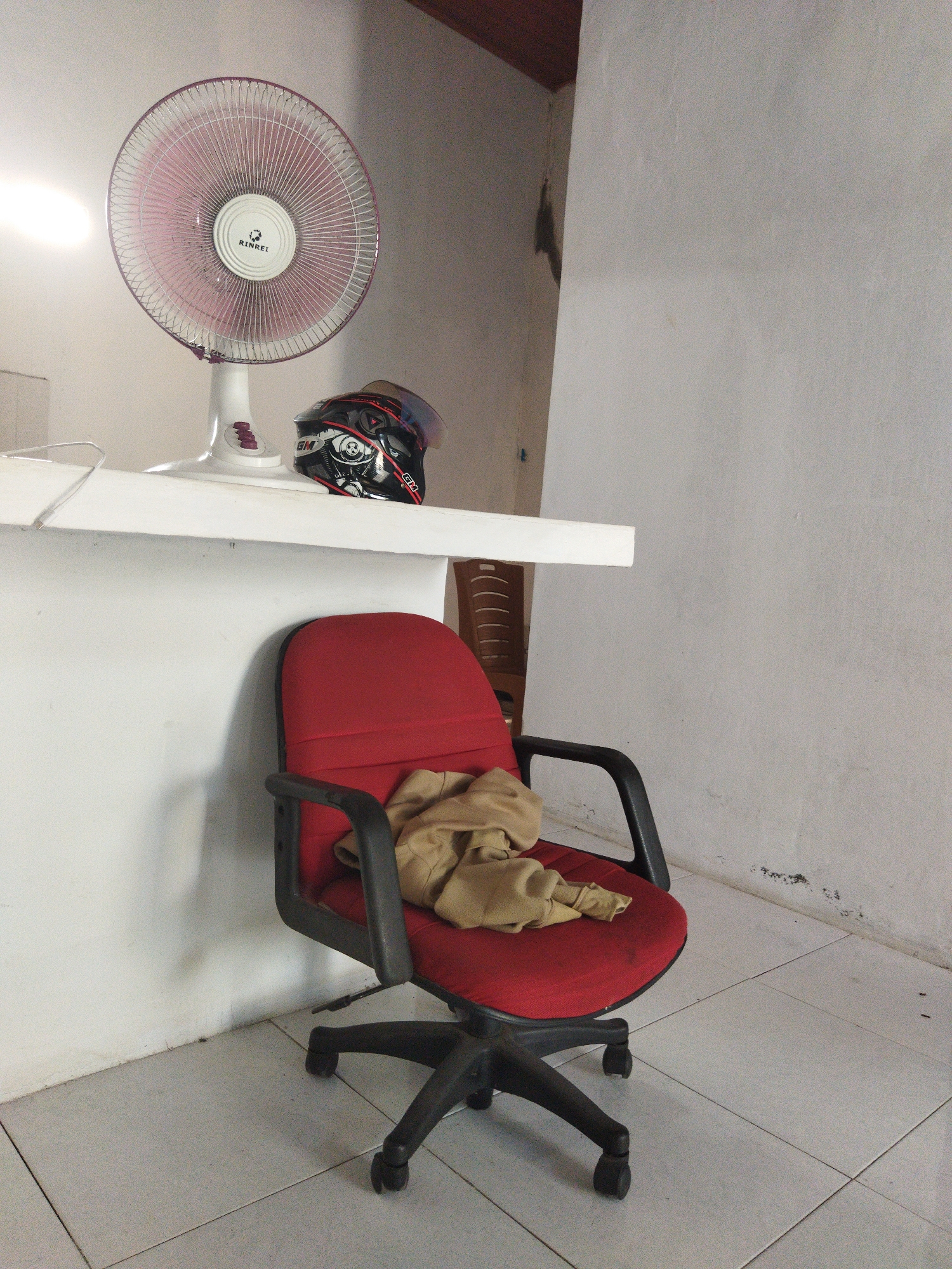 An office chair with a tan garment draped over its seat is positioned in front of a white wall with some discoloration. To the left of the chair, a white pedestal fan with a pink grill is situated on a white shelf, next to a black and red motorcycle helmet. The floor is tiled in a light gray color. The scene is likely indoors, possibly in a residential or small commercial space, and there are no people visible in the image. The overall impression is one of a common, perhaps utilitarian, interior space.