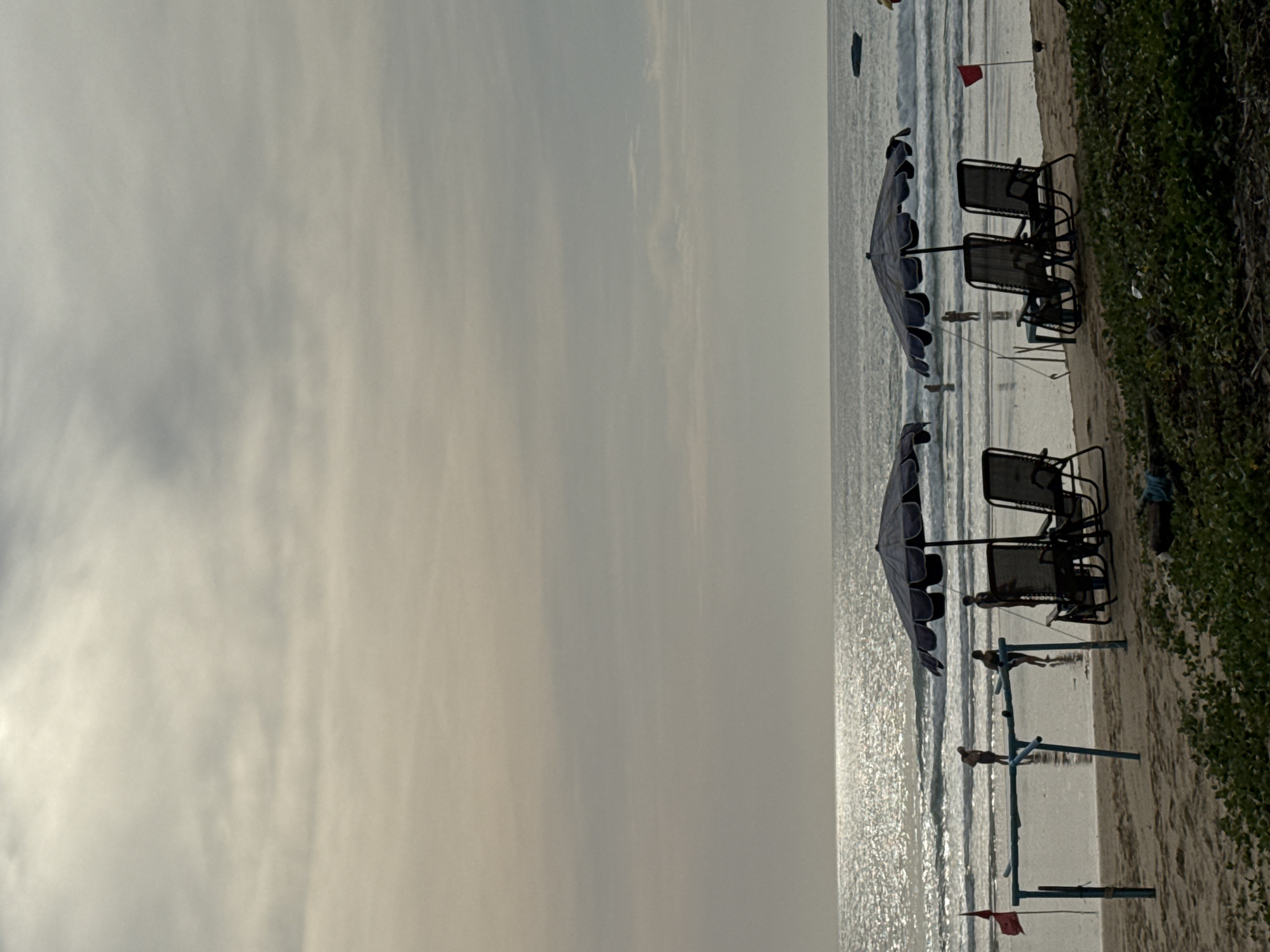 This is a vertical, eye-level shot of a beach scene in Karon, Thailand. The sky is overcast with muted gray clouds that diffuse the sunlight, creating a soft, ethereal glow on the ocean's surface. The ocean itself is a calm, choppy expanse with gentle waves rolling onto the shore, reflecting the bright light from the sky.
On the sandy beach, two dark blue umbrellas stand sentinel over two pairs of empty lounge chairs. The chairs are made of dark mesh fabric stretched over metal frames, and they are positioned to face the ocean. A red flag flutters on a thin pole to the left of the chairs, and another red flag is visible further down the beach on the right.
In the shallow water, several people are visible. Two figures, one male and one female, are standing near the water's edge, appearing to be wading or playing in the surf. Another person, partially obscured by a blue structure, is also in the water. In the distance, a dark boat is visible on the horizon.
The foreground of the image is dominated by lush, green vegetation, some of which appears to be ground cover and other plants with larger leaves. The sand is a light tan color, with some debris scattered across it. The overall atmosphere is one of peaceful tranquility, with the natural beauty of the beach setting enhanced by the gentle weather.