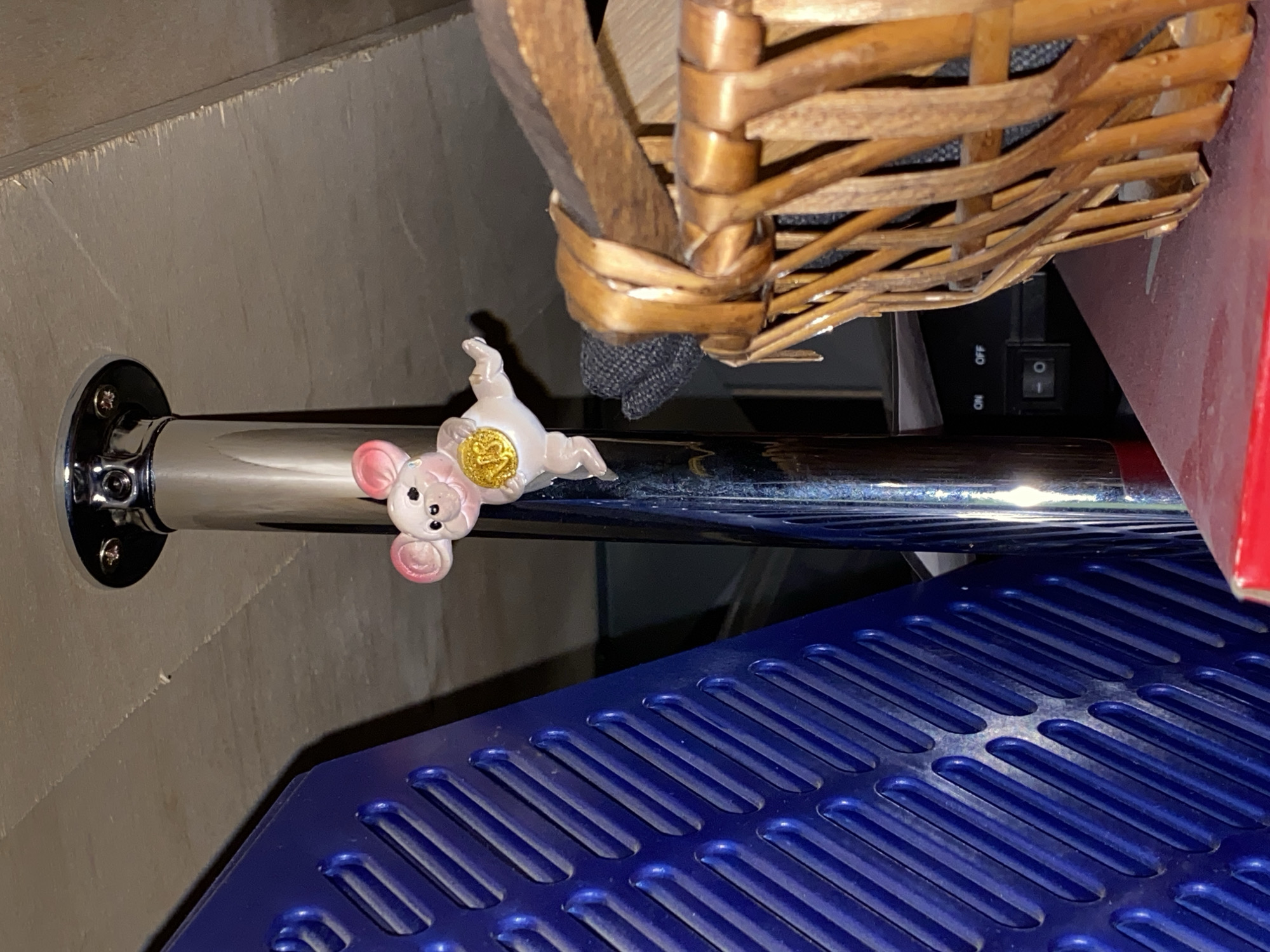 A small, pink, plastic figurine of a mouse is affixed to a vertical chrome pole. The mouse is positioned sideways and appears to be reaching out with one hand, holding a gold coin with the number "18" embossed on it. The pole is attached to a circular chrome fixture at the top, which is screwed into a light-colored, possibly wooden, surface. To the left of the pole is a bright blue object with a textured, ribbed surface. To the right and in the foreground, a portion of a woven basket made of light brown material is visible. The background is dark and out of focus, with hints of electronic equipment and a red surface visible at the bottom. The image is taken from a low angle, looking upwards. This scene does not provide any specific details to determine a location in the city of Moskva, Russia.